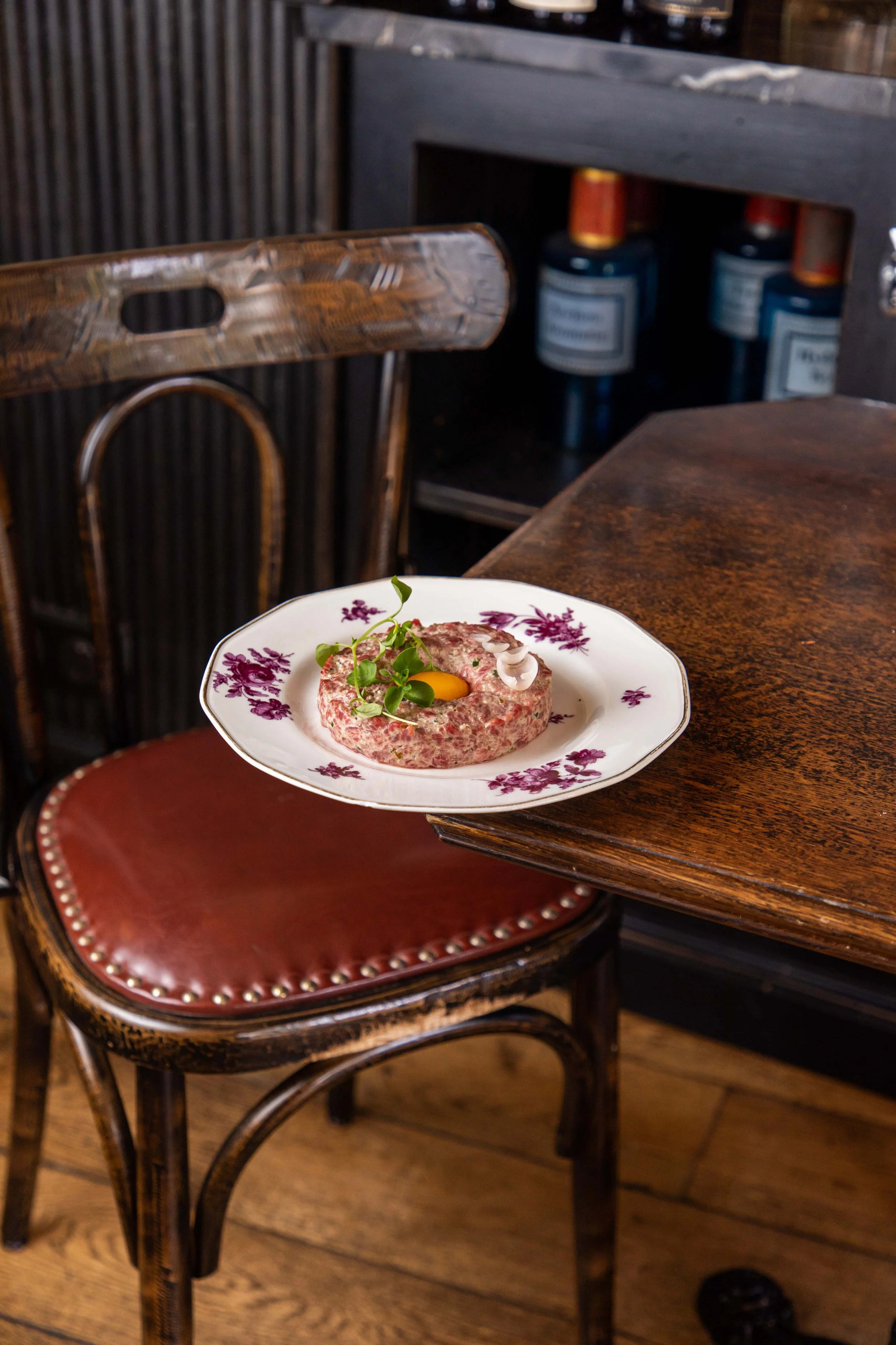 Une assiette avec un tartare de viande, garnie d'une tranche d'œuf cru, de microgreens et de copeaux de fromage, posée sur une chaise en bois avec un siège en cuir marron. En arrière-plan, on voit une table en bois et des bouteilles sur une étagère.
