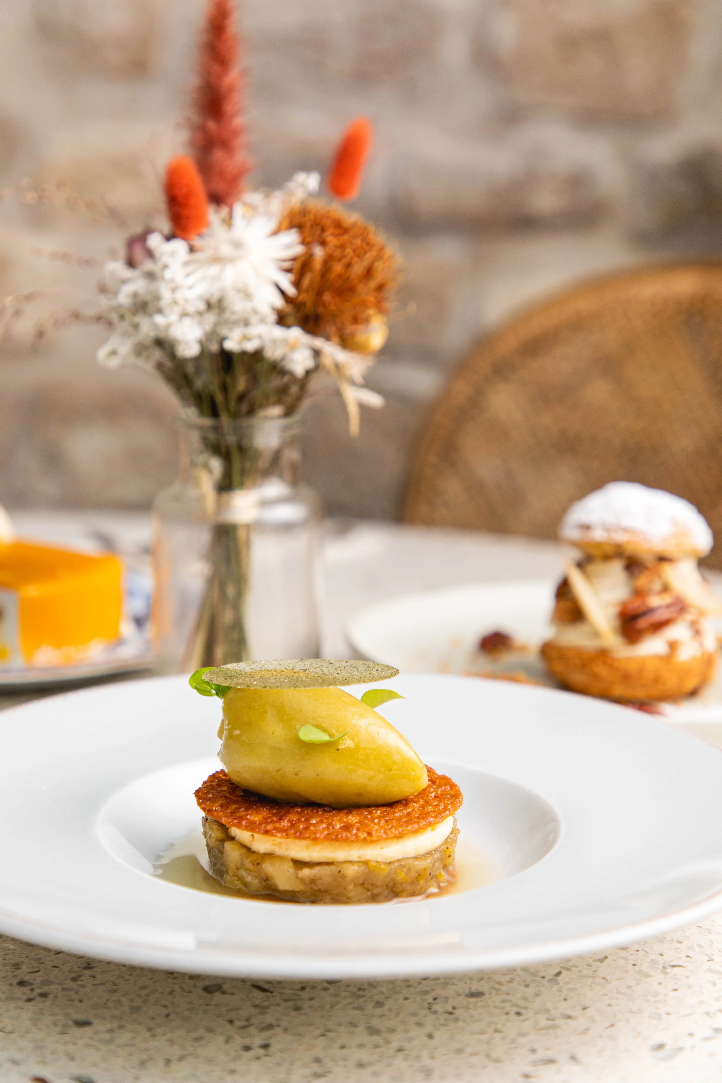Une assiette avec un plat gastronomique, comprenant une pièce de viande ou poisson, une purée ou sauce, une quenelle de mousse ou de crème et une fine tranche de biscuit ou croustille par-dessus, sur une table avec un bouquet de fleurs et un dessert 