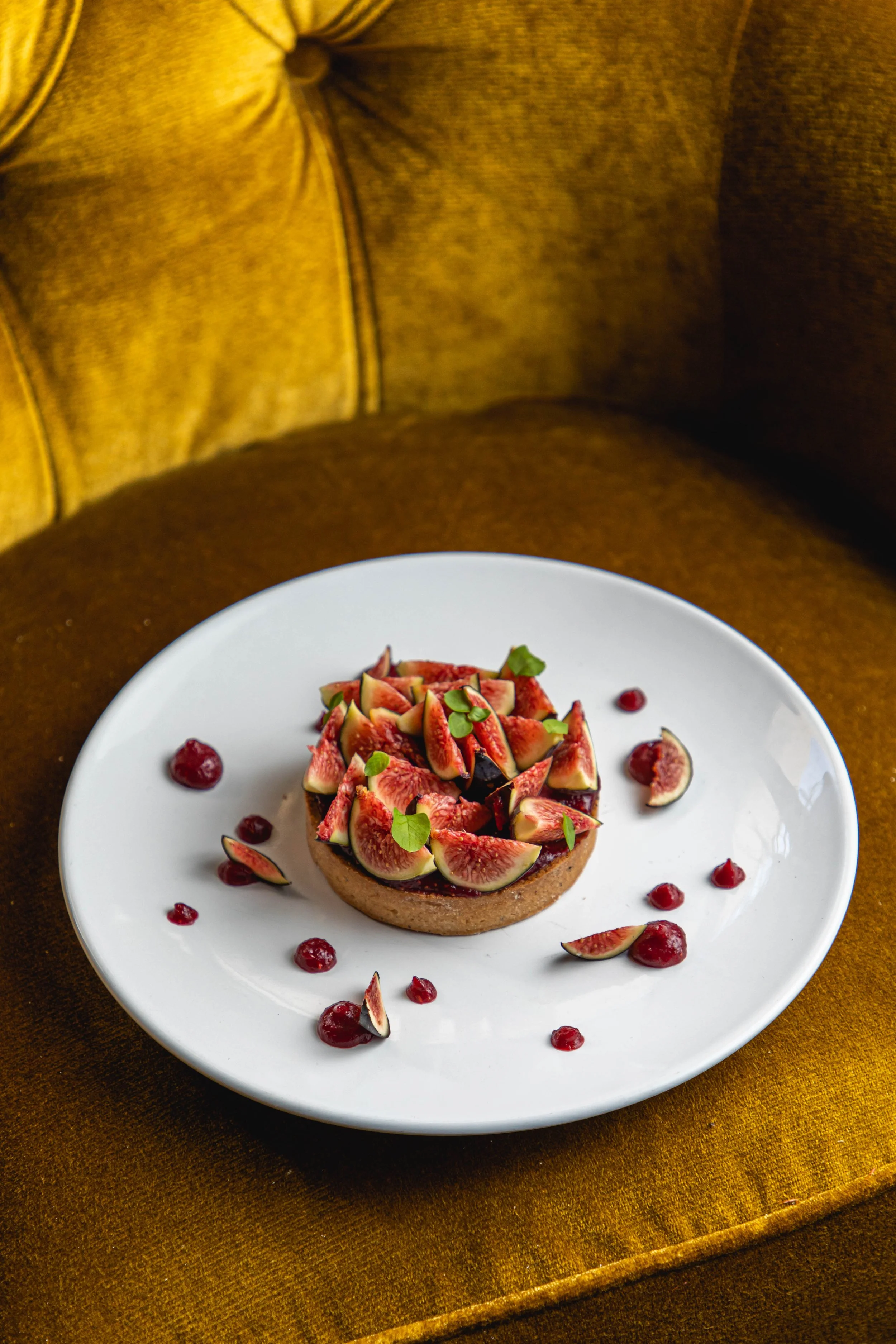Tarte aux figues fraîches garnie de feuilles vertes sur une assiette blanche, avec des morceaux de figues et des graines de grenade autour, sur un fauteuil en velours marron.