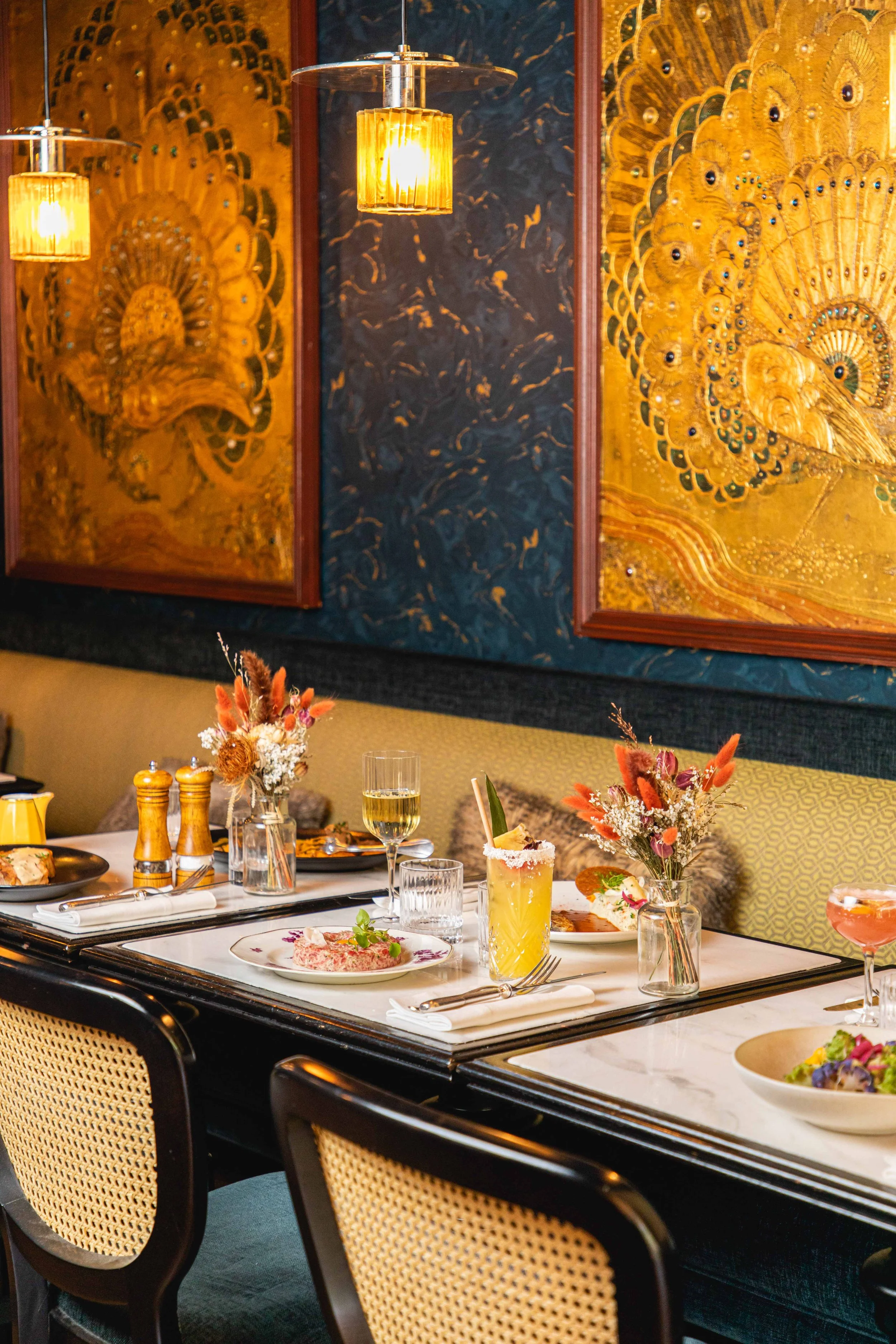 Une table de restaurant élégante avec des plats, des boissons et des bouquets de fleurs, sur fond de mur décoratif avec des peintures de plumes de paon dorées, éclairée par un éclairage chaud.