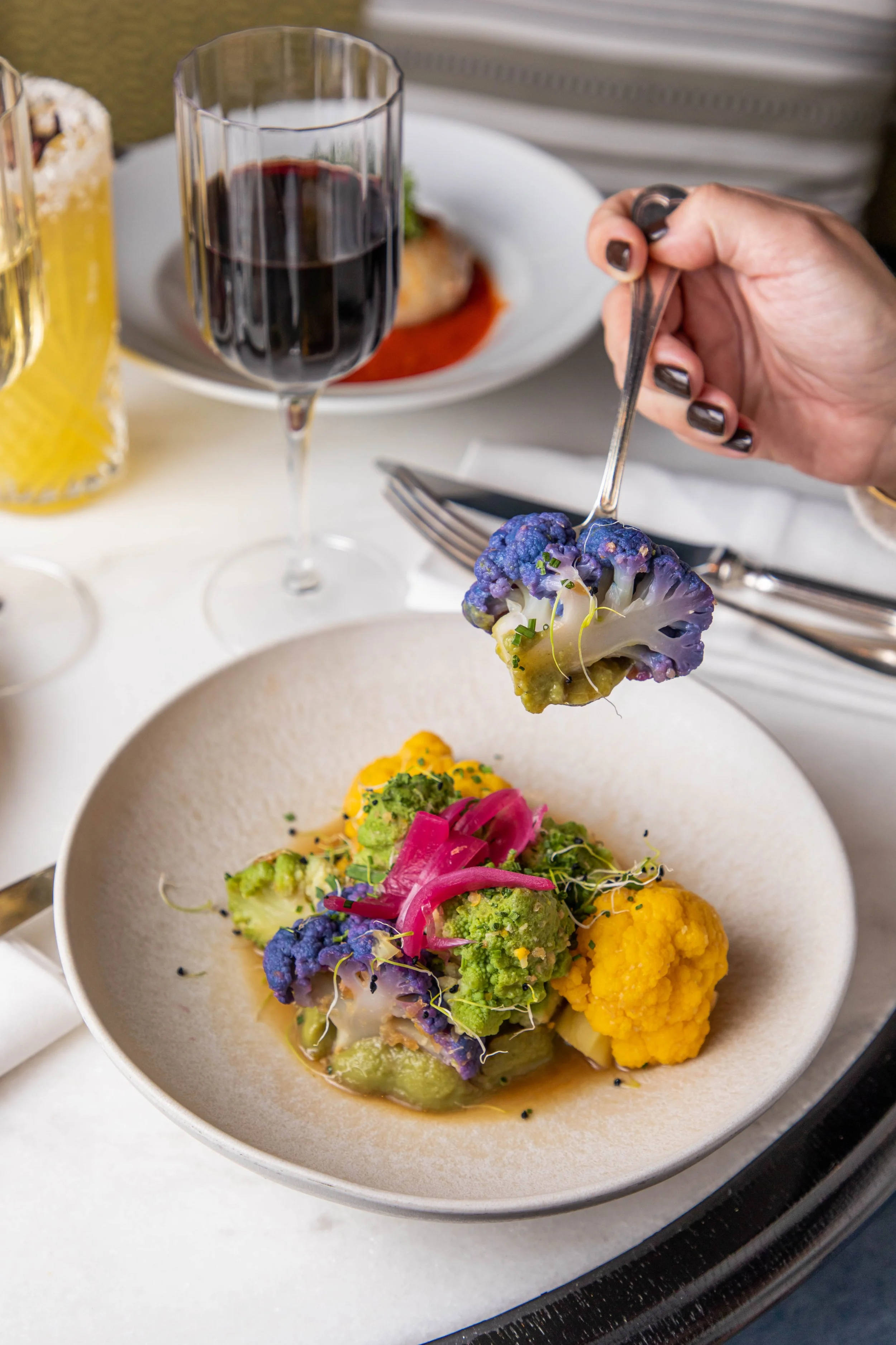 Assiette de légumes colorés, notamment chou-fleur violet, vert et jaune, avec des oignons roses, sur une table de restaurant, avec verre de vin rouge et autre assiette en arrière-plan.