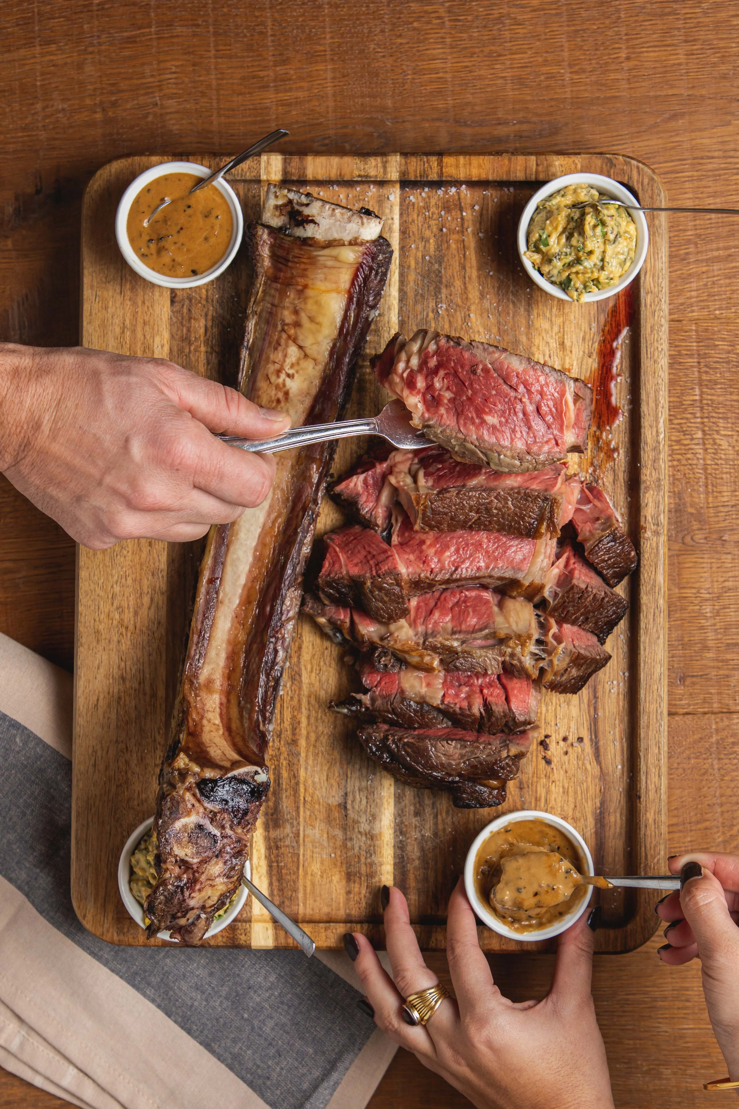 Un plateau de viande de bœuf, côtes de bœuf épaisses, avec des sauces et des accompagnements dans de petits bols. Deux mains utilisent des fourchettes pour couper et servir la viande.