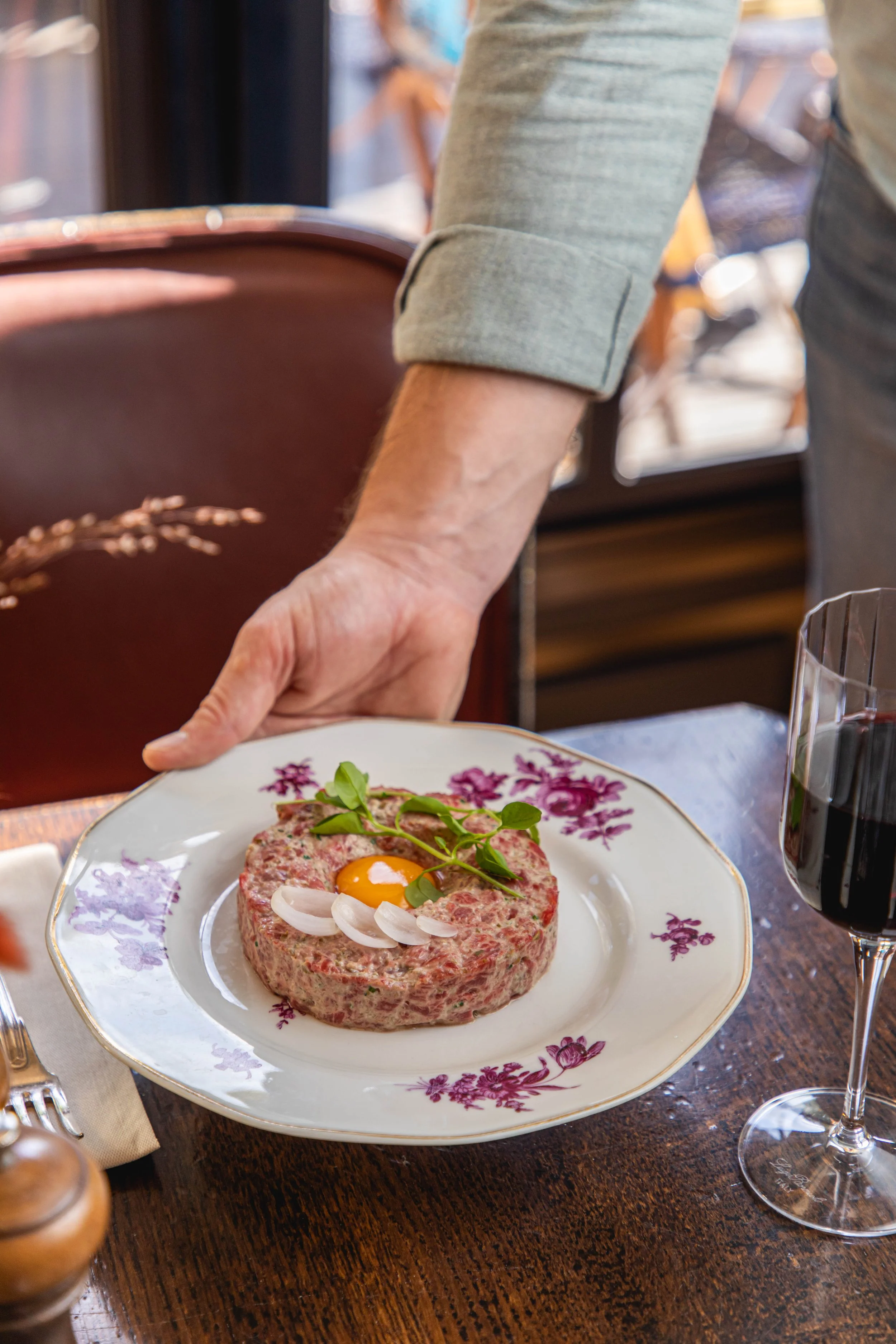 Un homme sert un tartare de viande avec un jaune d'œuf cru, décoré de feuilles vertes et de tranches d'oignon, sur une assiette blanche à motifs violets, dans un restaurant.
