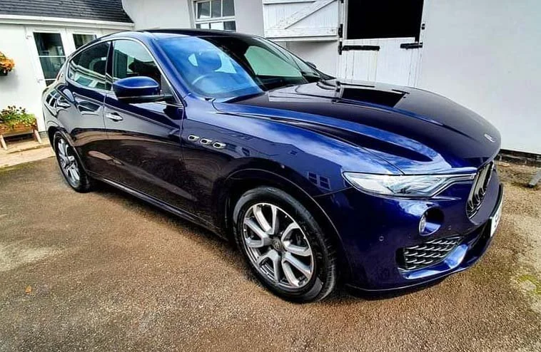 A dark blue sedan parked on a driveway near a white house with black shutters.