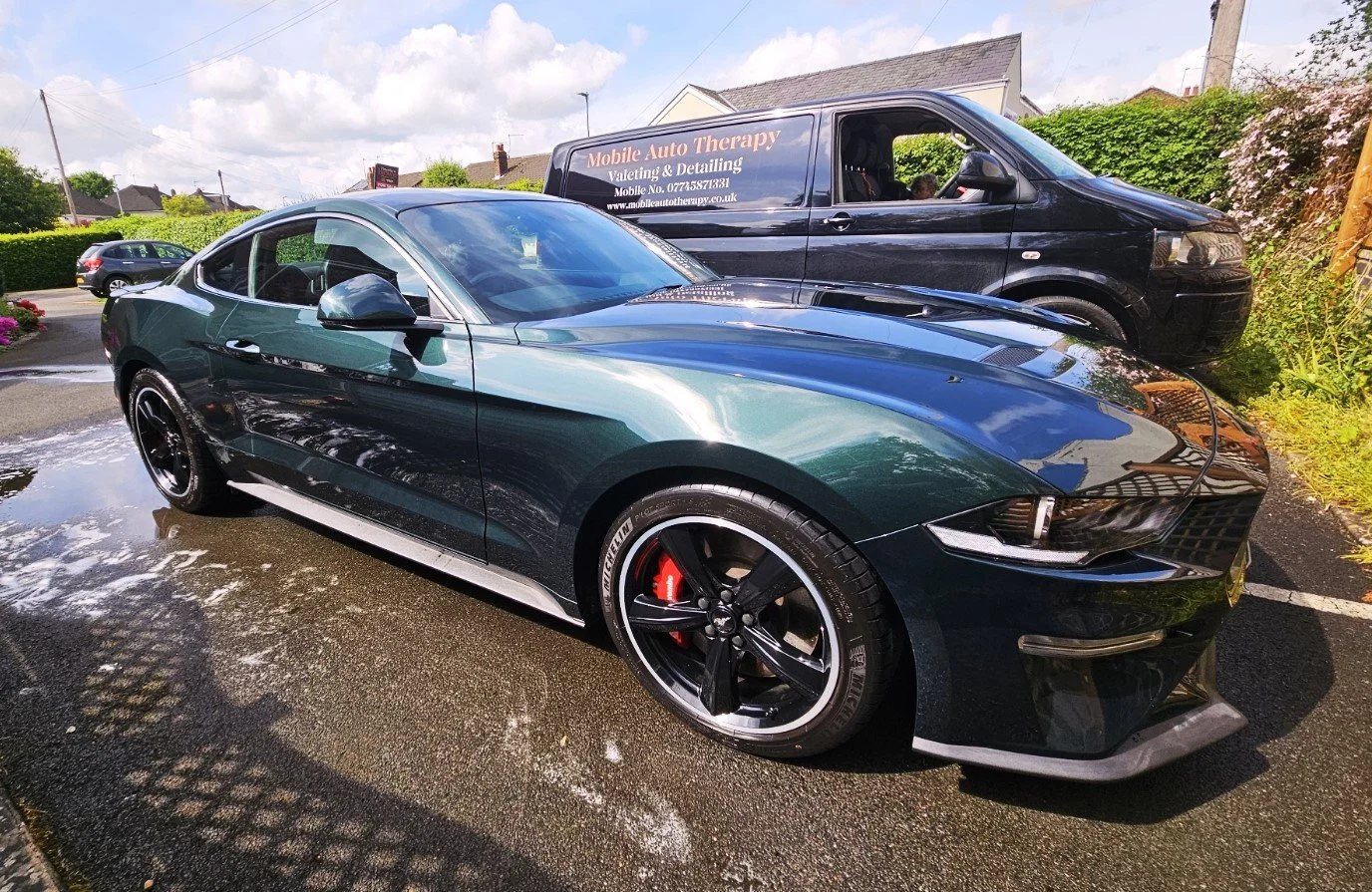 A black sports car parked on wet pavement, with a black van behind it. The sports car has black wheels with red brake calipers and is shiny, reflecting the sky and surroundings.