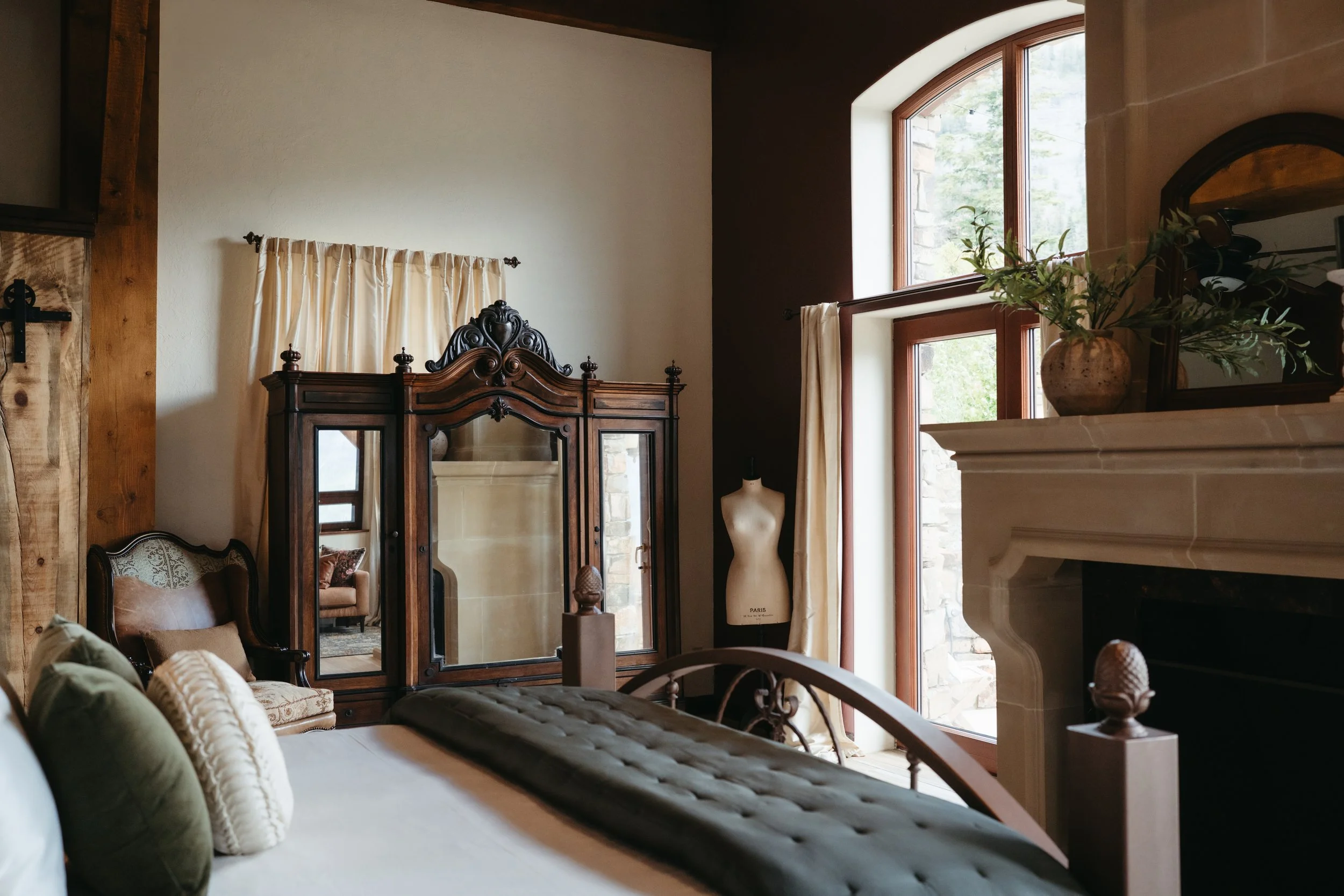 A cozy bedroom with a bed, decorative pillows, a vintage wooden wardrobe, a mannequin, a fireplace with a plant on the mantel, and large arched windows.