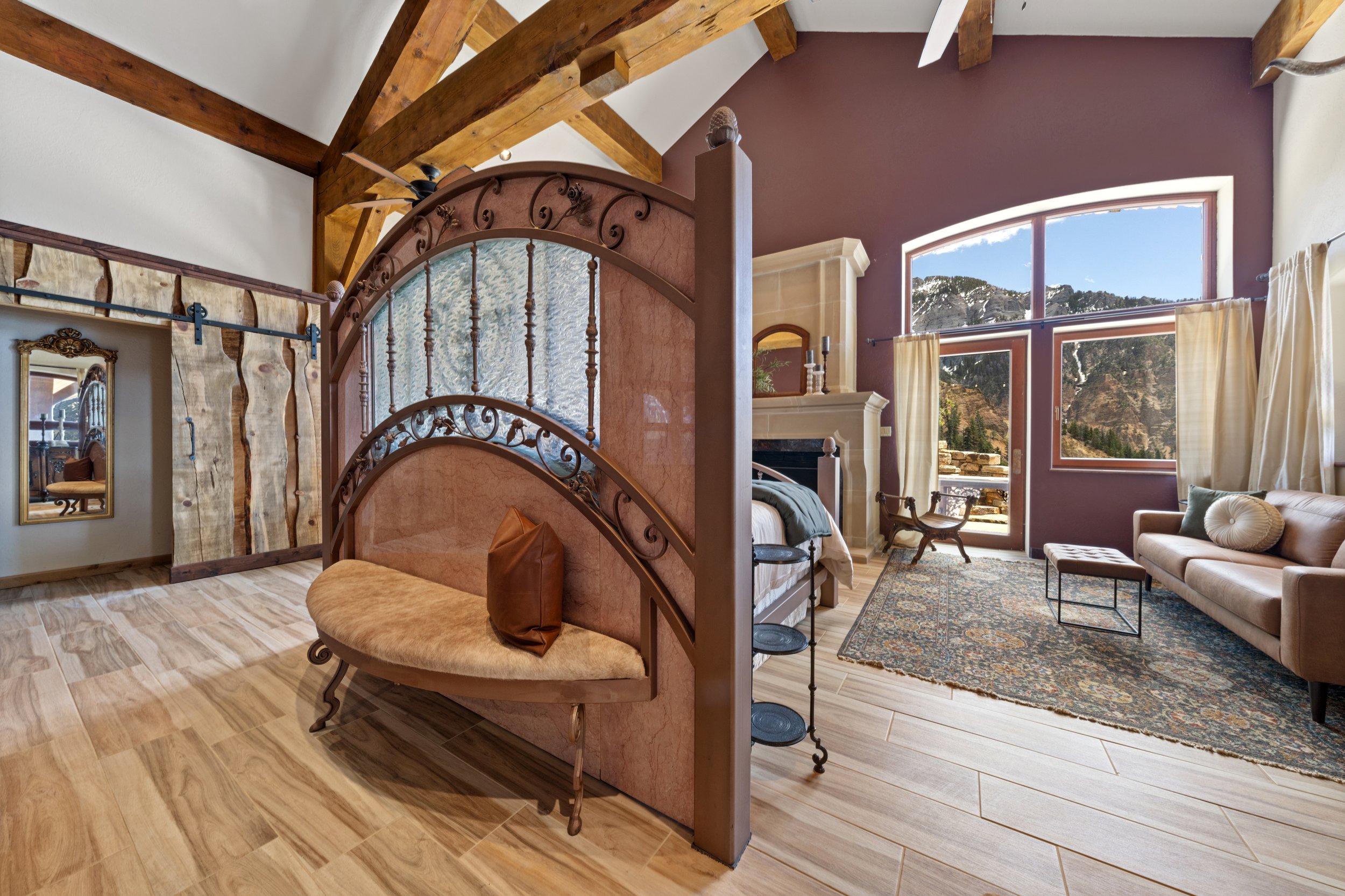 Spacious bedroom with wood beams, large windows showing mountain view, fireplace, beige sofa, area rug, and a partially visible bed with a headboard.