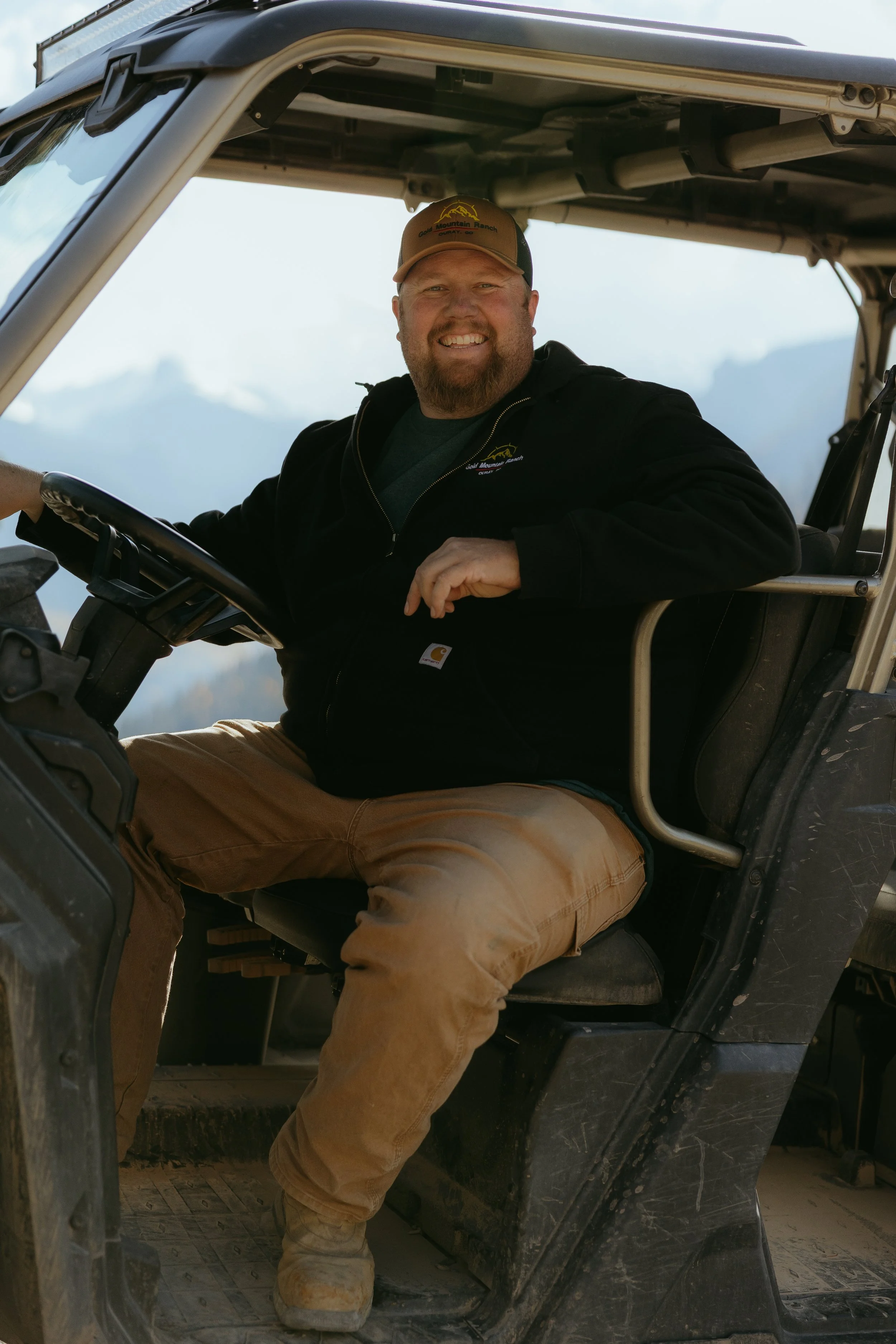 A smiling man sitting in the driver's seat of a utility vehicle, wearing a black jacket, tan work pants, boots, and a baseball cap with a mountain/logo design.