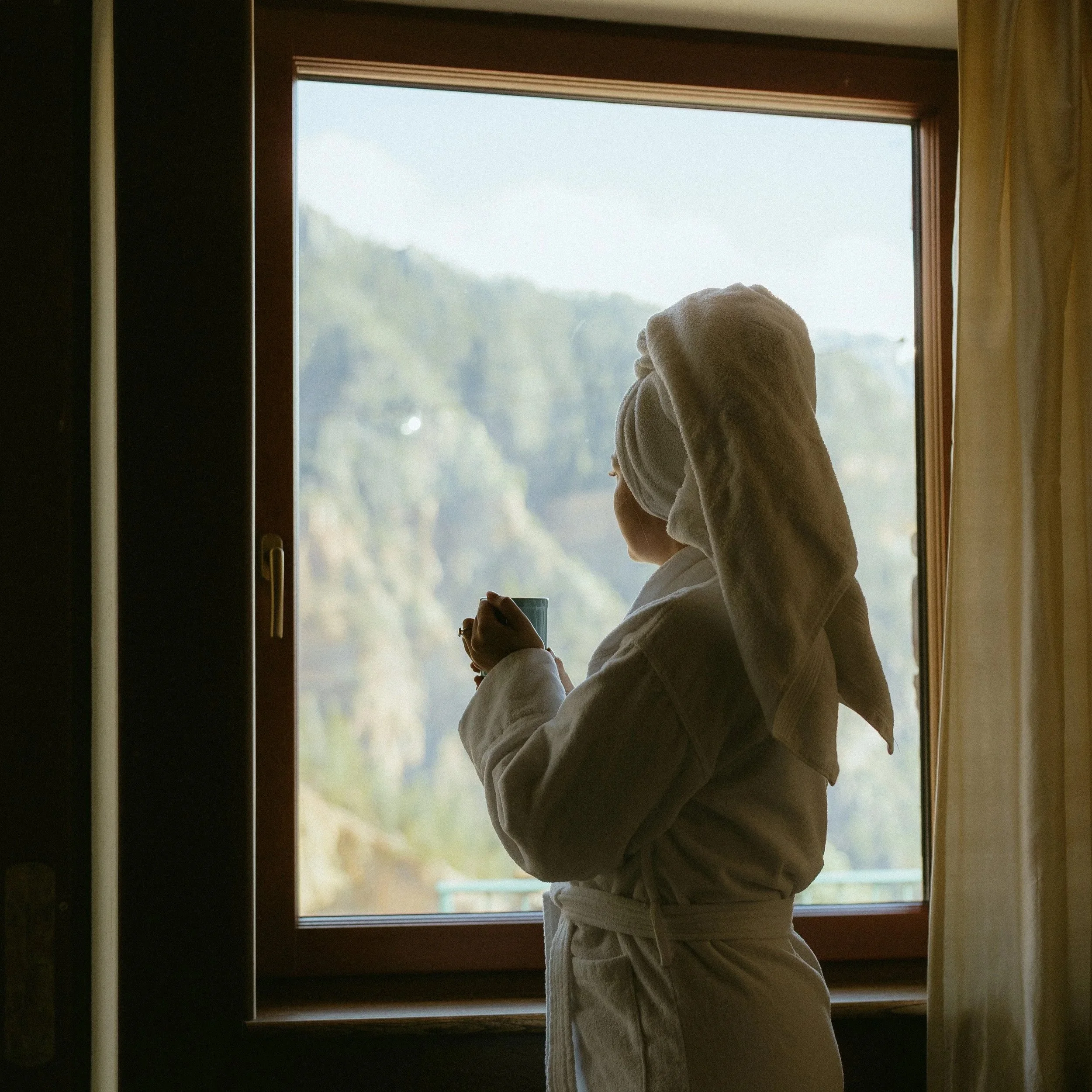 A person wearing a bathrobe and towel on their head standing by a window, holding a cup, looking outside at scenic mountains.