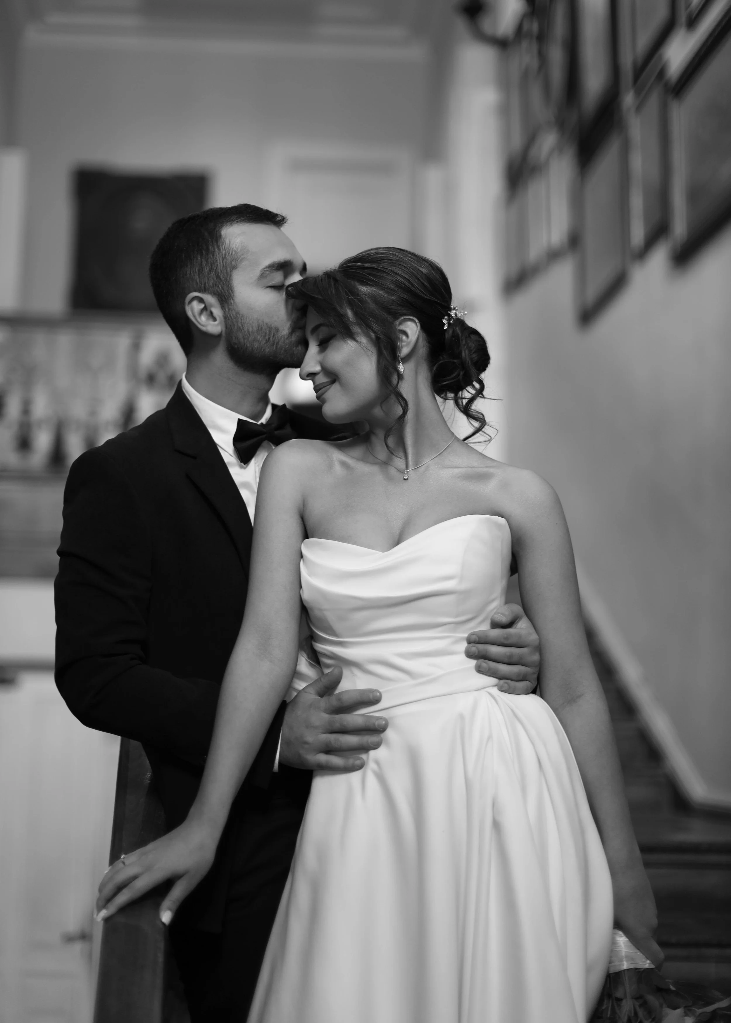 Black and white photo of a newlywed couple, the groom with short dark hair and beard, wearing a tuxedo with bow tie, kissing the bride's forehead as she smiles, wearing a strapless wedding dress with her hair in an elegant updo, in a room with framed photos on the wall.