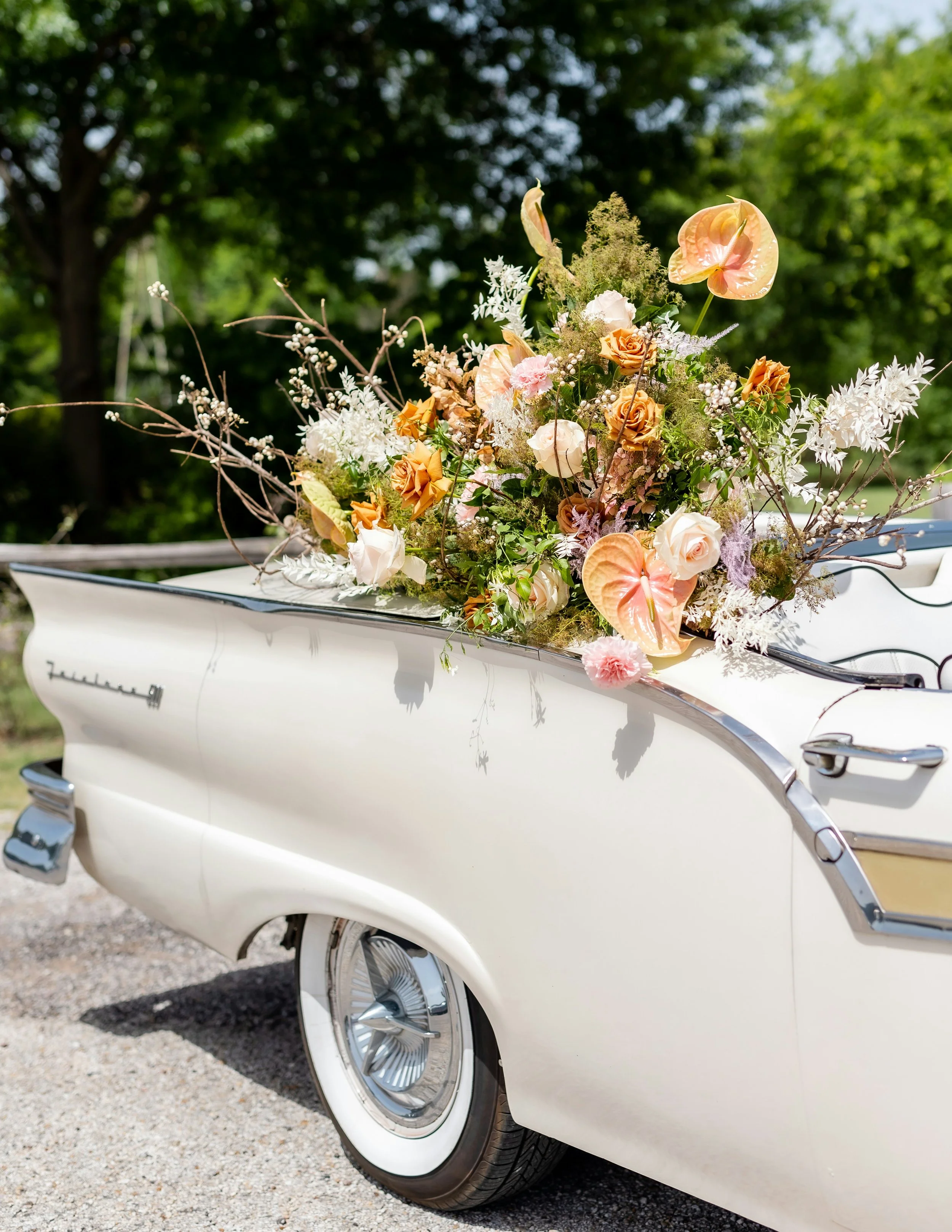 A vintage white car decorated with a large floral arrangement on its trunk, featuring various flowers and greenery.