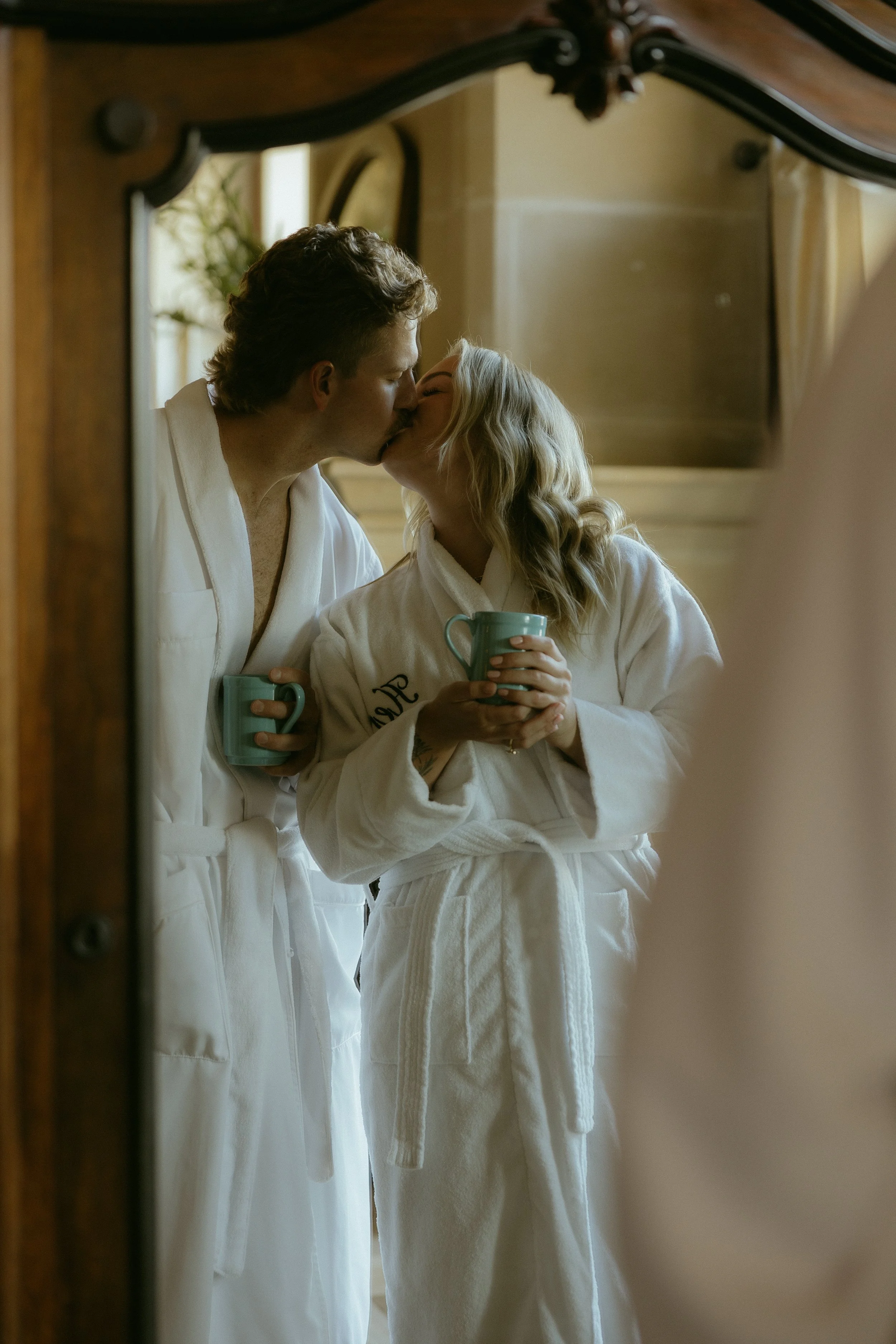 A couple in white bathrobes sharing a kiss while holding coffee mugs, reflected in a mirror in a cozy, warmly lit room.