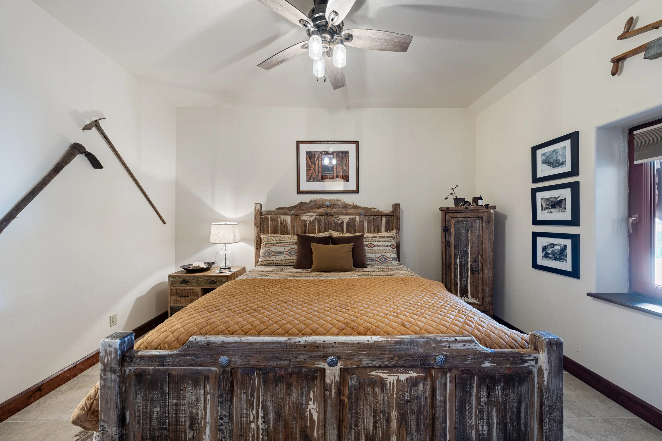 Bedroom with rustic wooden bed, orange quilt, decorative pillows, framed pictures on the walls, window with wooden frame, ceiling fan with lights, wooden furniture, and wall-mounted fencing tools.
