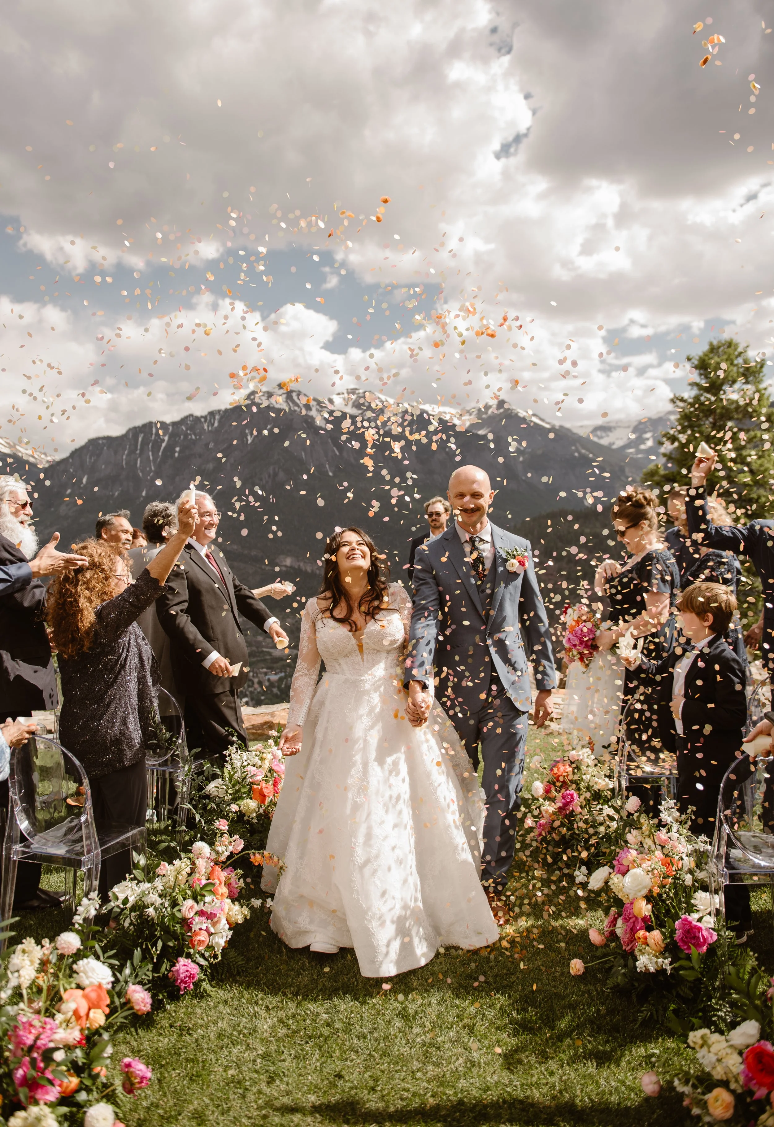A bride and groom walking hand-in-hand through a confetti celebration outdoors with mountains in the background.