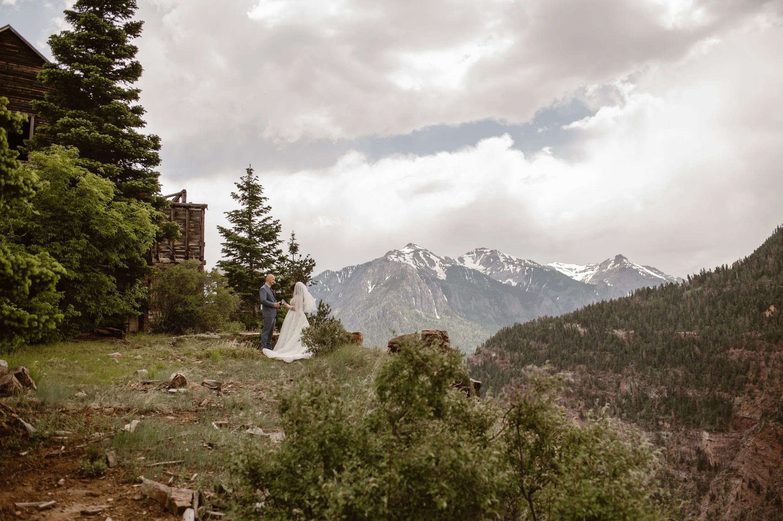 A wedding ceremony taking place outdoors on a mountain backdrop with snow-capped peaks, a bride and groom facing each other, surrounded by trees and cloudy sky.