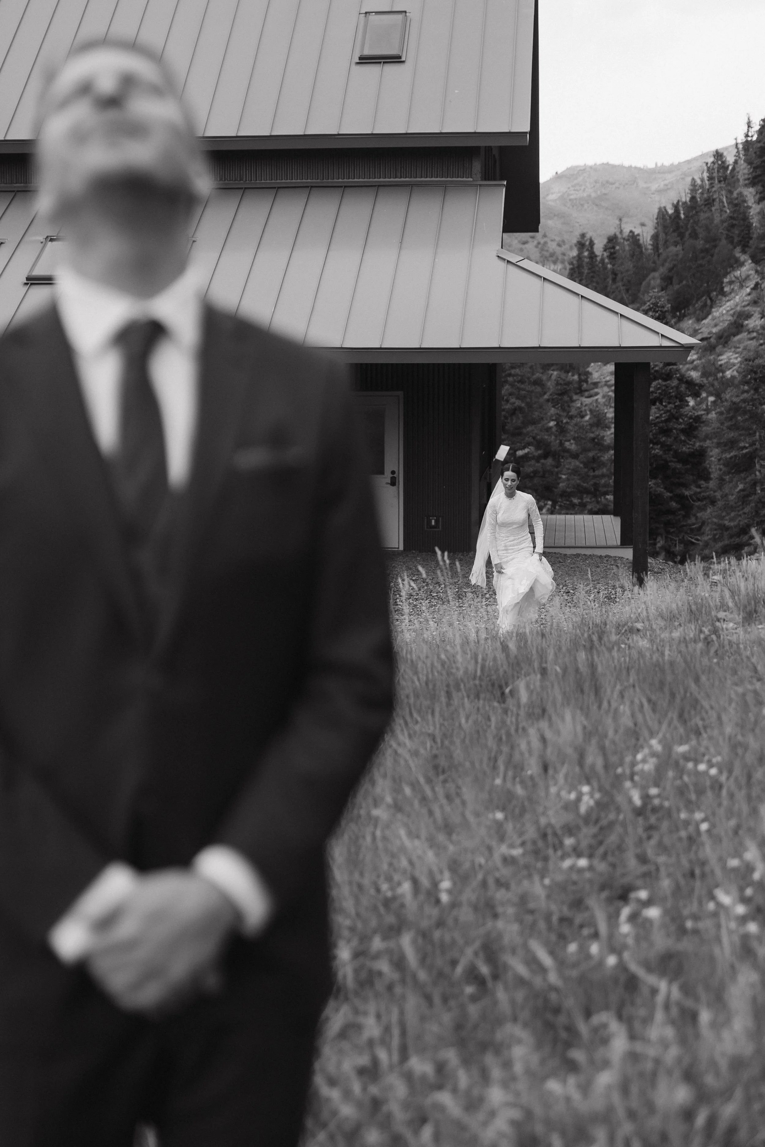 A man in a suit standing in the foreground, slightly out of focus, with a woman in a wedding dress holding a bouquet of flowers walking toward him in the background in an outdoor setting with tall grass and a house.