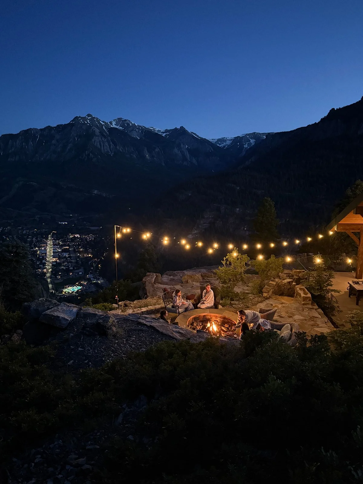 People sitting around a fire pit on a mountain terrace at night, with string lights above and a view of a mountain town below.