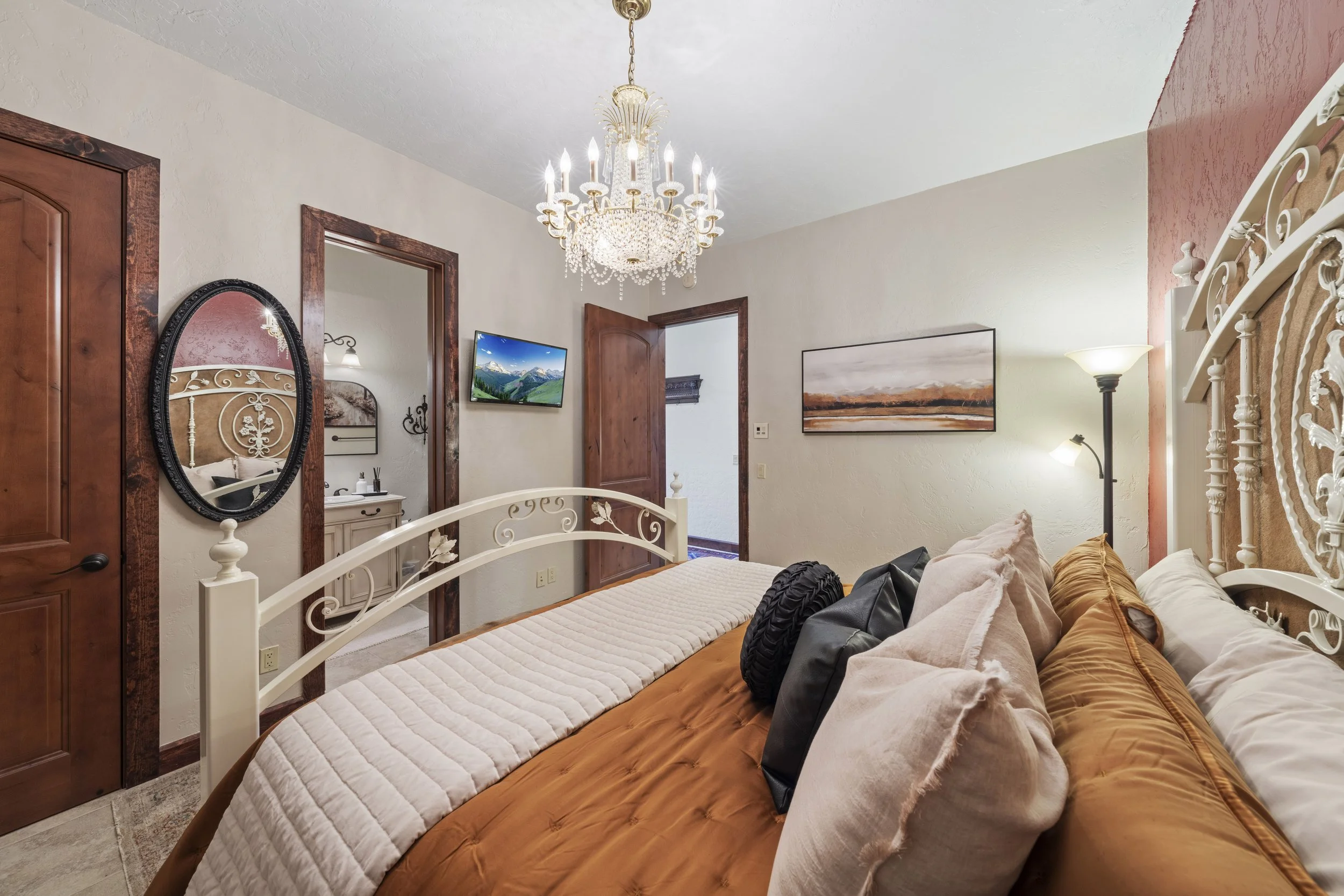 Bedroom with a bed, pillows, a chandelier, a wall mirror, a TV, doorways, and artworks on the walls