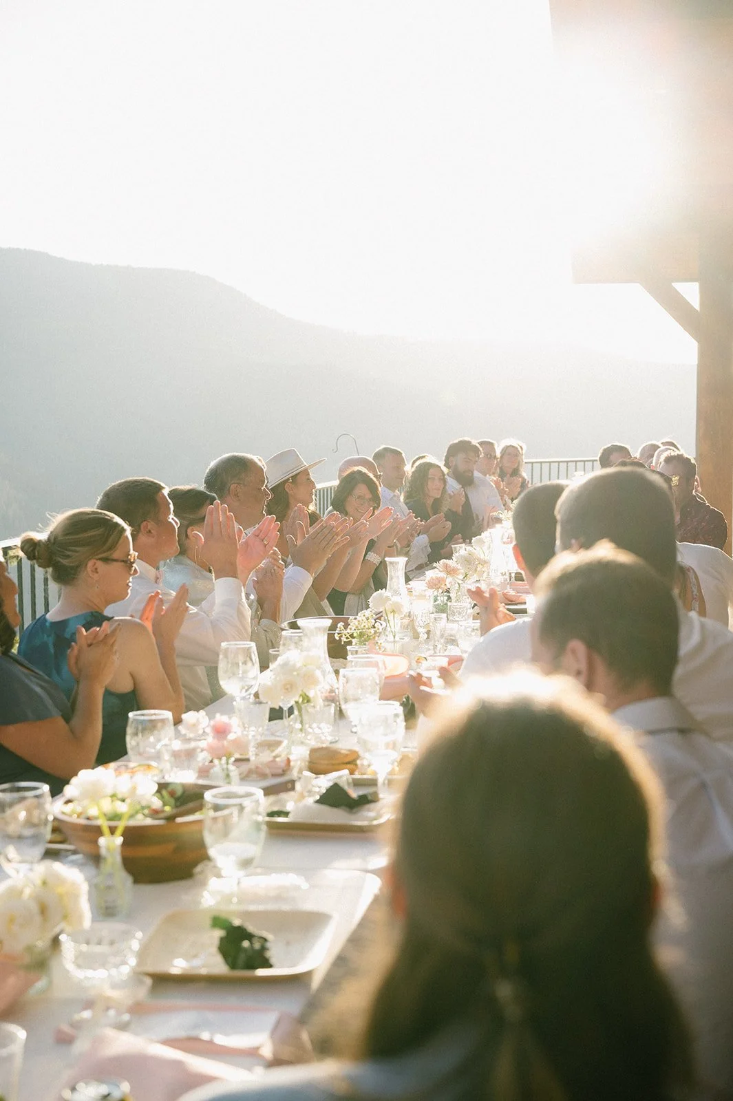 People gathered around a long table celebrating outdoors at sunset, clapping, with mountains in the background.