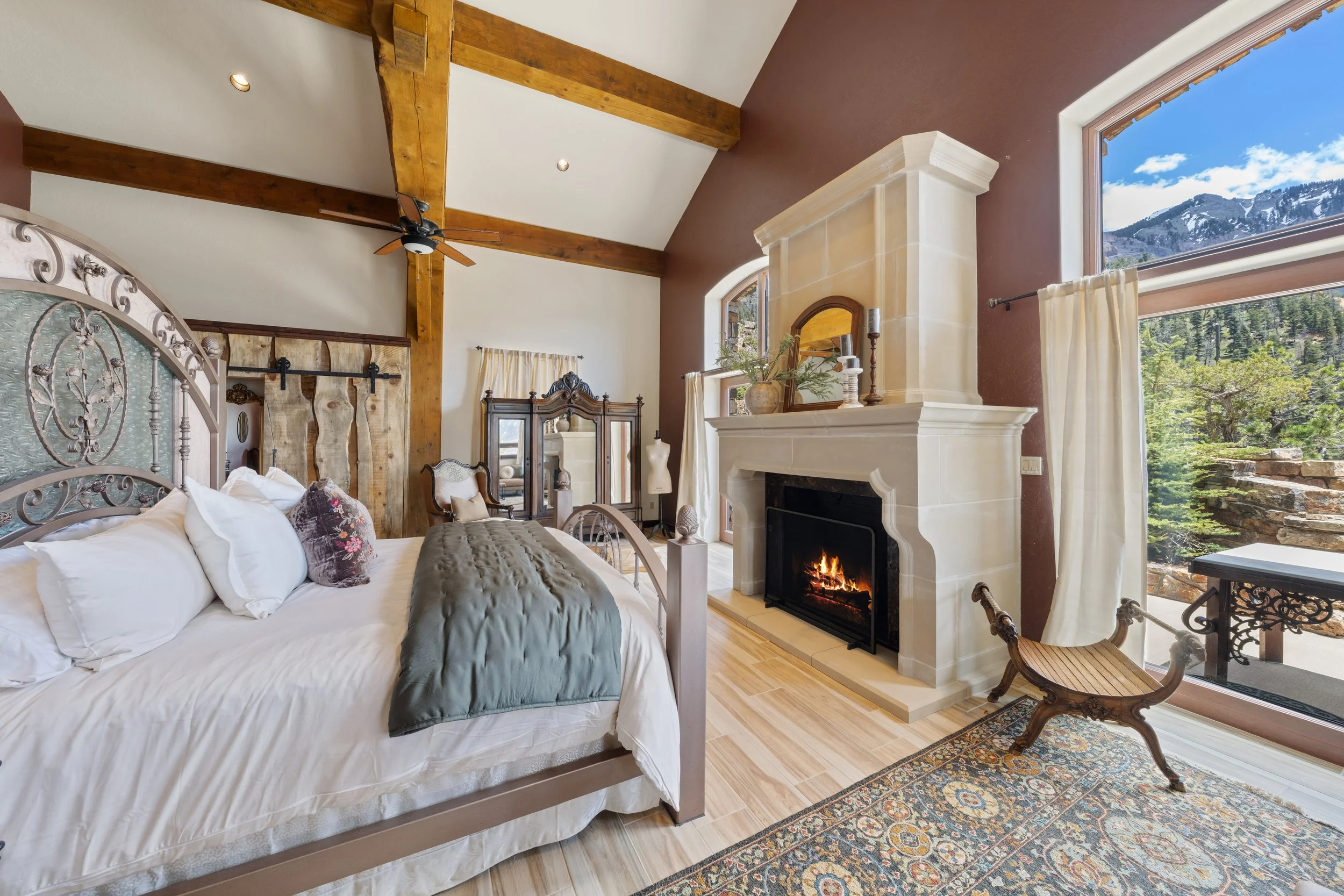 Interior of a bedroom featuring a white bed with pillows, a fireplace with a fire, large windows showing mountains and trees, and wooden beams on the ceiling.