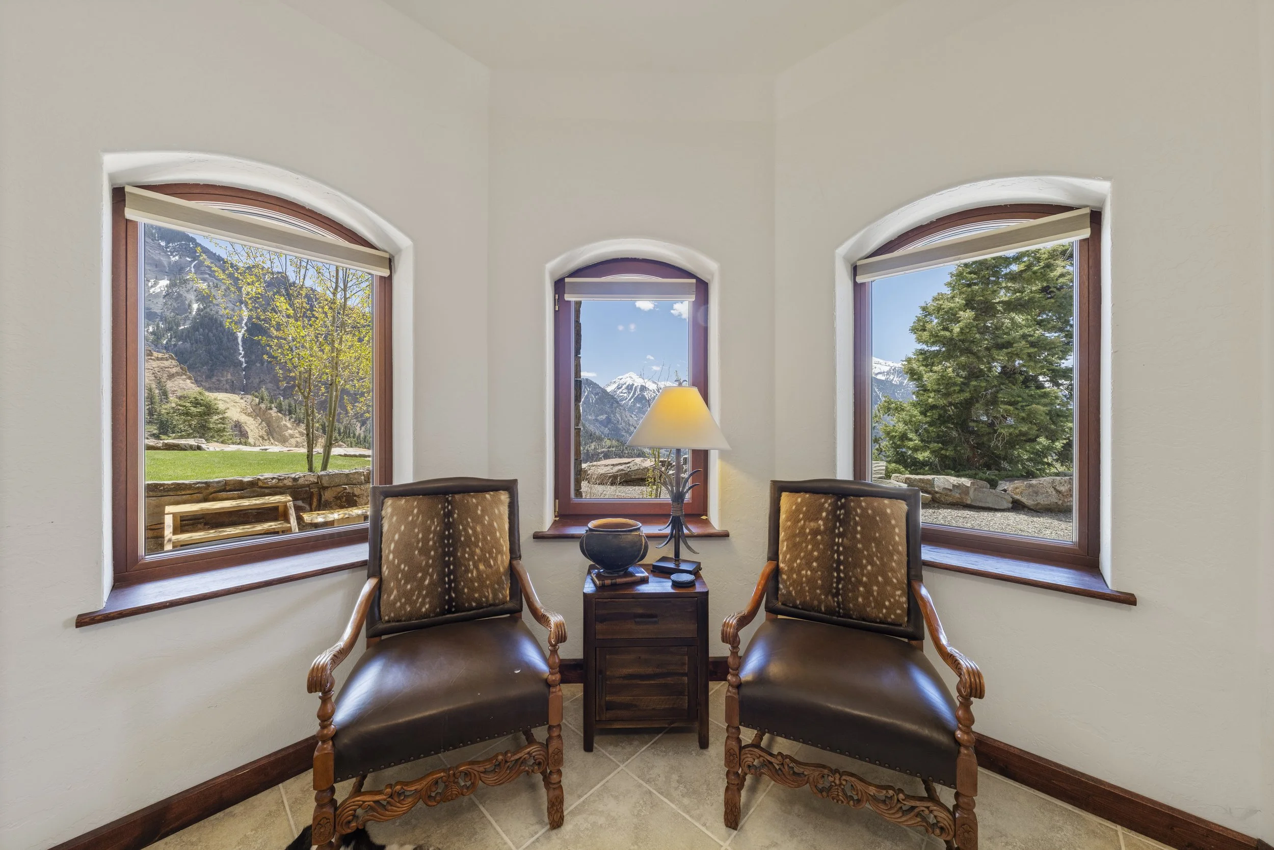A cozy sitting area with two wooden chairs with padded seats and backrests, placed on either side of a small wooden side table with a lamp, set against a background of three large windows showcasing mountainous scenery and trees.