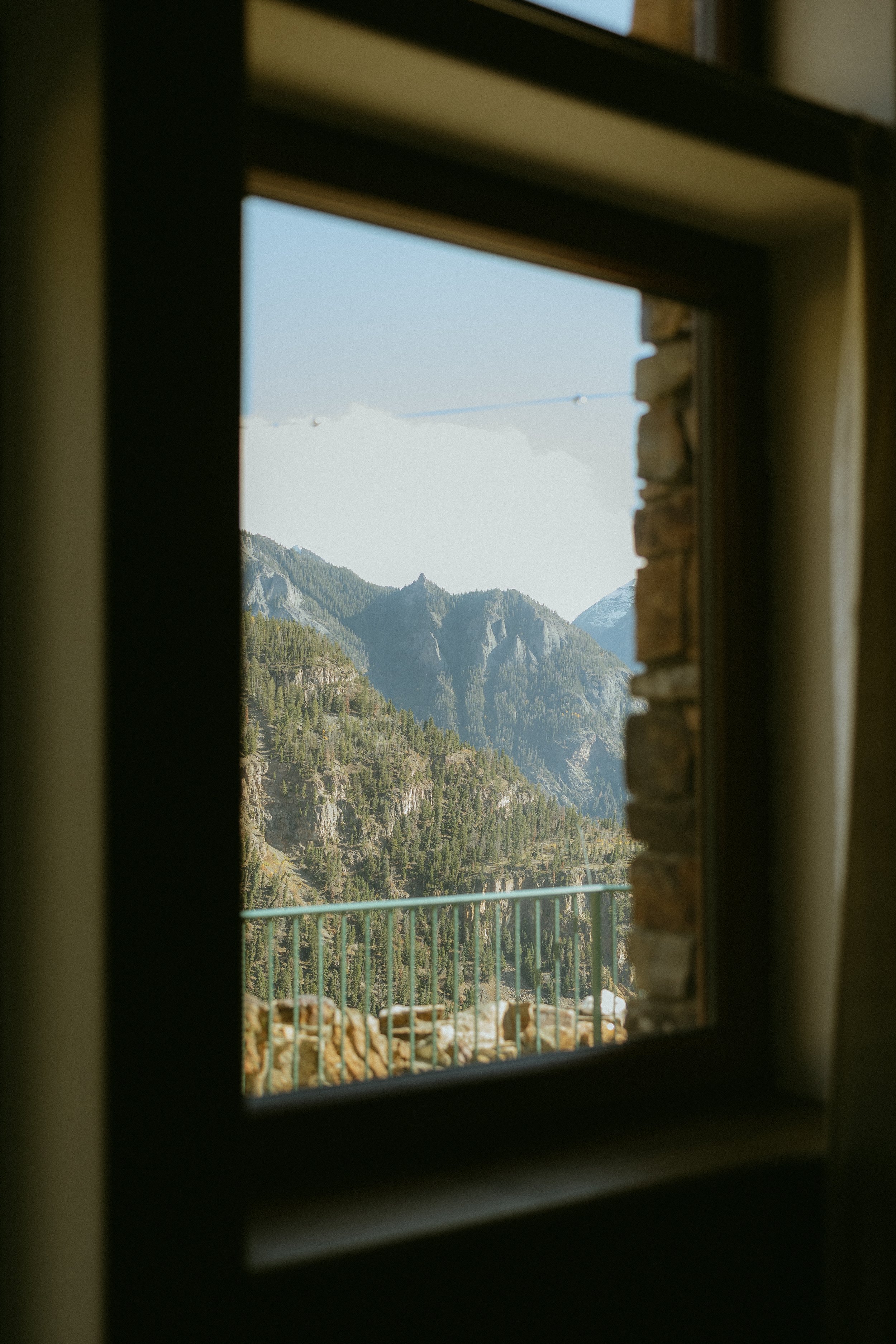 View of a mountain landscape through a window, with green pine trees and rocky peaks under a clear blue sky.