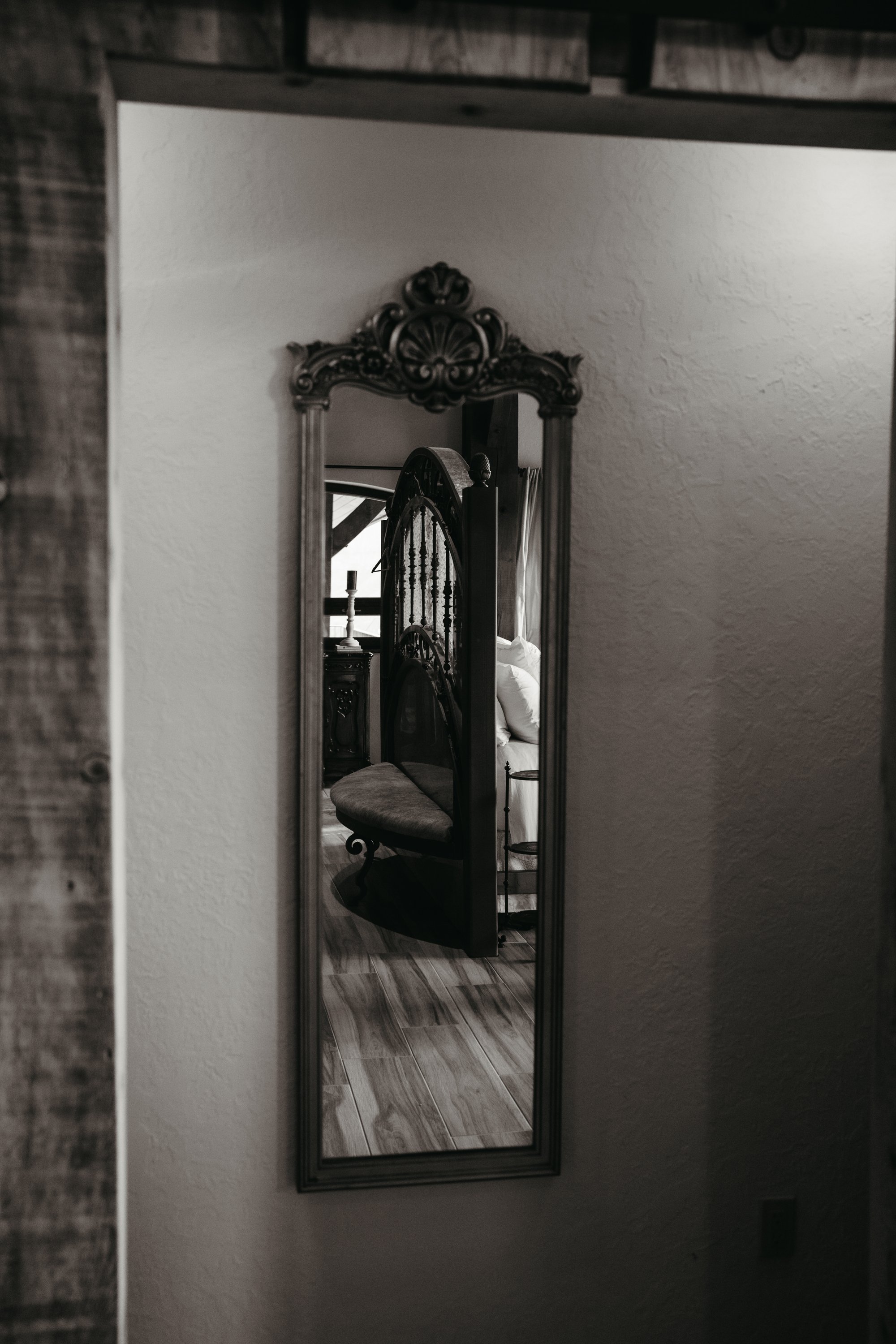 A narrow mirror with an ornate wooden frame reflects a bedroom with a vintage-style metal bed, pillows, a side table, and a candle holder.