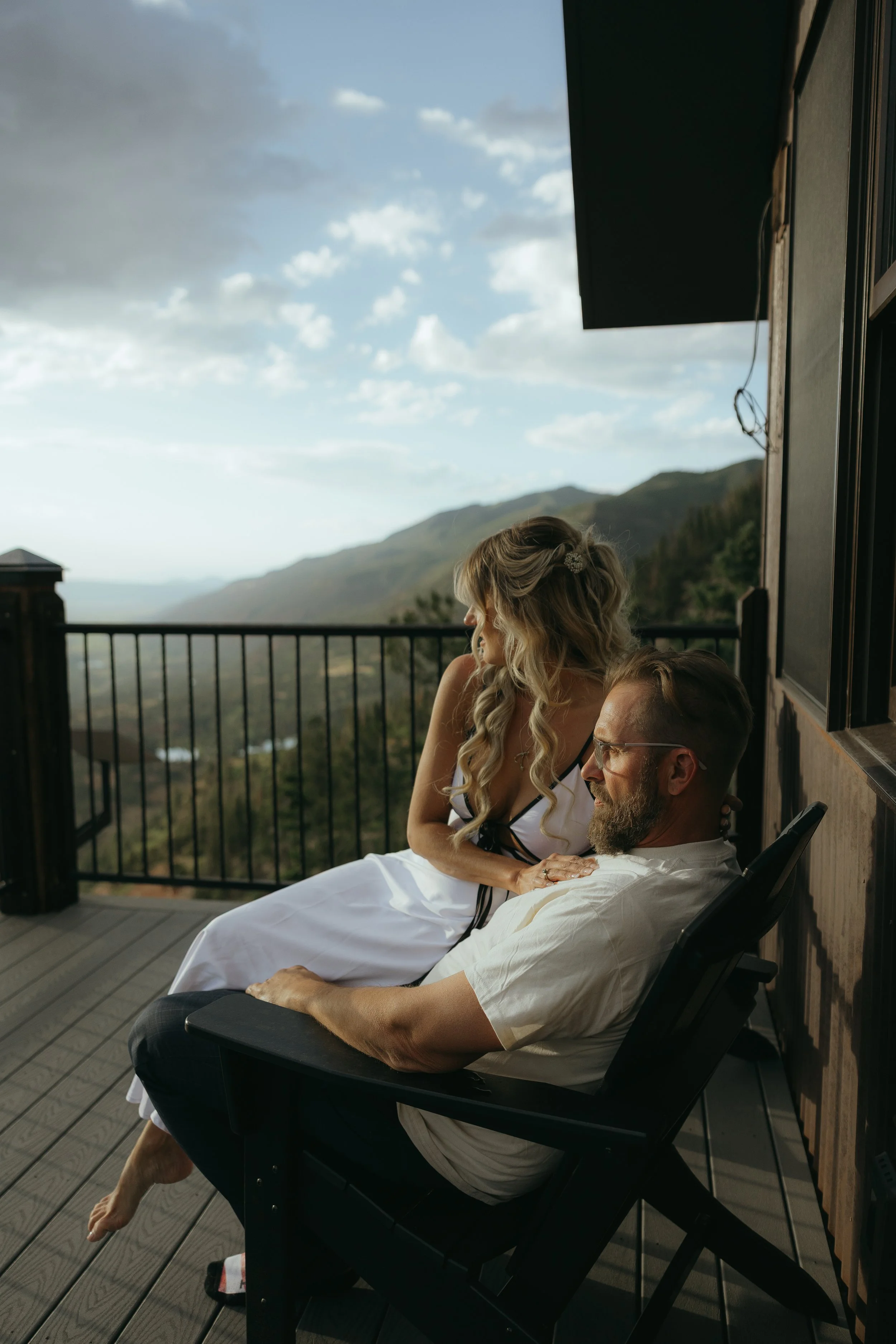 A man and woman sitting on a deck with scenic mountain view, the woman resting her hand on the man's shoulder.