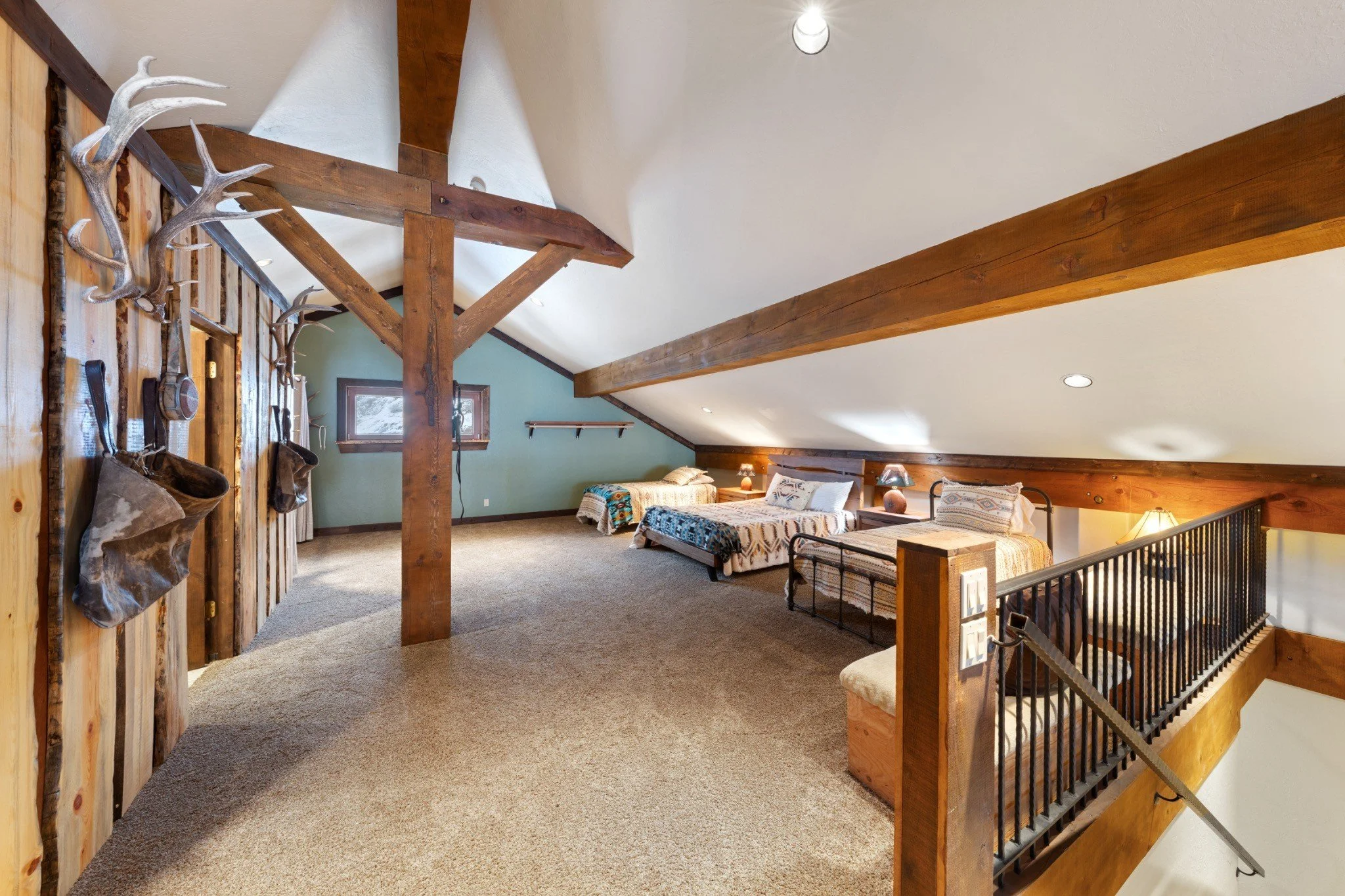 A spacious attic bedroom with three beds, wooden beams, a sloped ceiling, and rustic decor, including a mounted set of antlers and a wall with hanging leather and metal items.