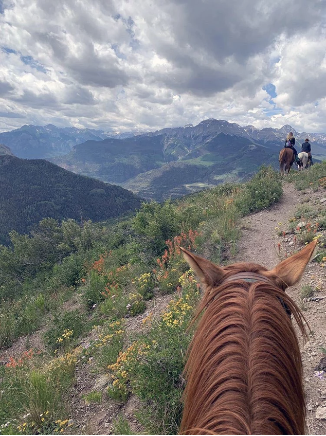 View from horseback riding trail showing two riders on horses walking along a grassy path with mountain range and cloudy sky in the background.