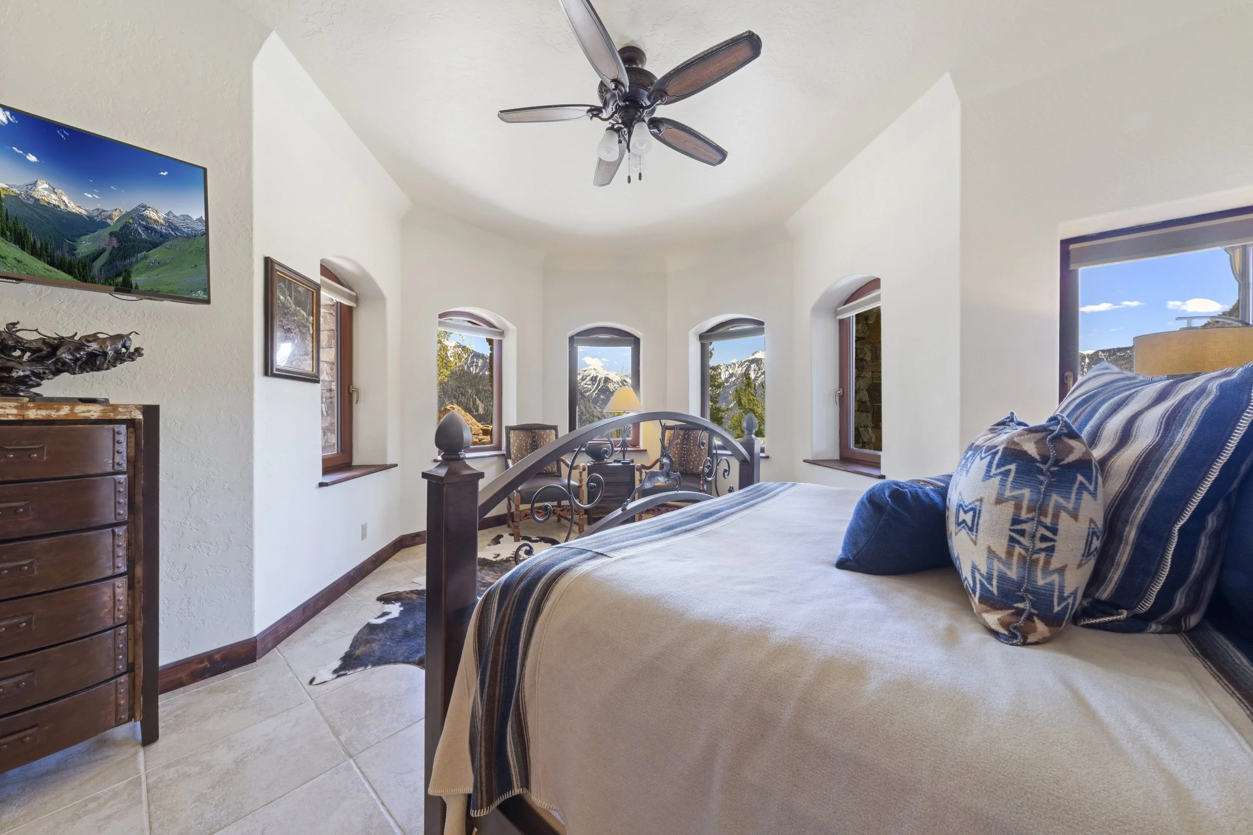 A bedroom with a large bed adorned with blue and beige striped pillows. Multiple windows showcase mountain scenery. A wooden dresser and a ceiling fan are also visible.