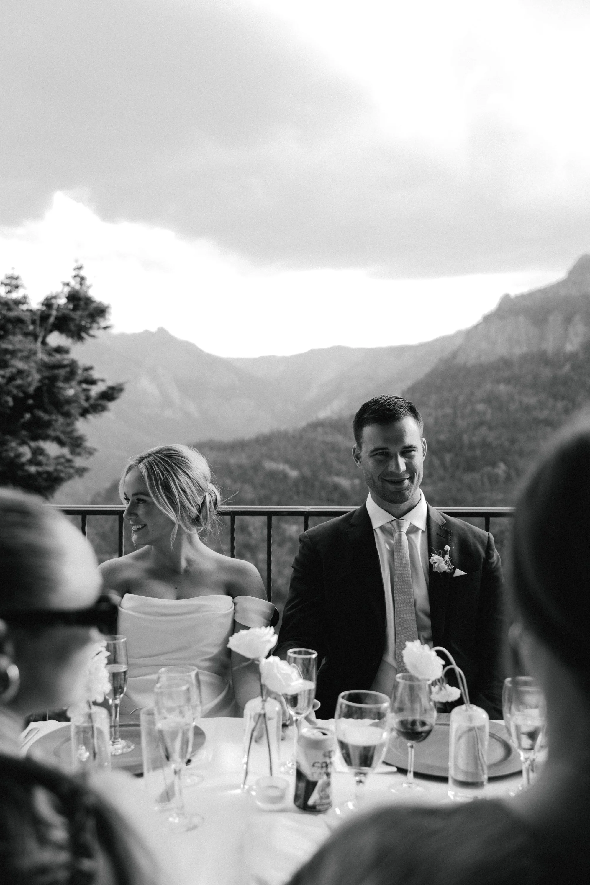 Black and white photo of wedding reception with a bride and groom sitting at a table outdoors with mountainous background. The bride has blonde hair and is smiling, while the groom, in a suit and tie, is smiling and looking forward. The table is set with glasses, flowers, and wine bottles.