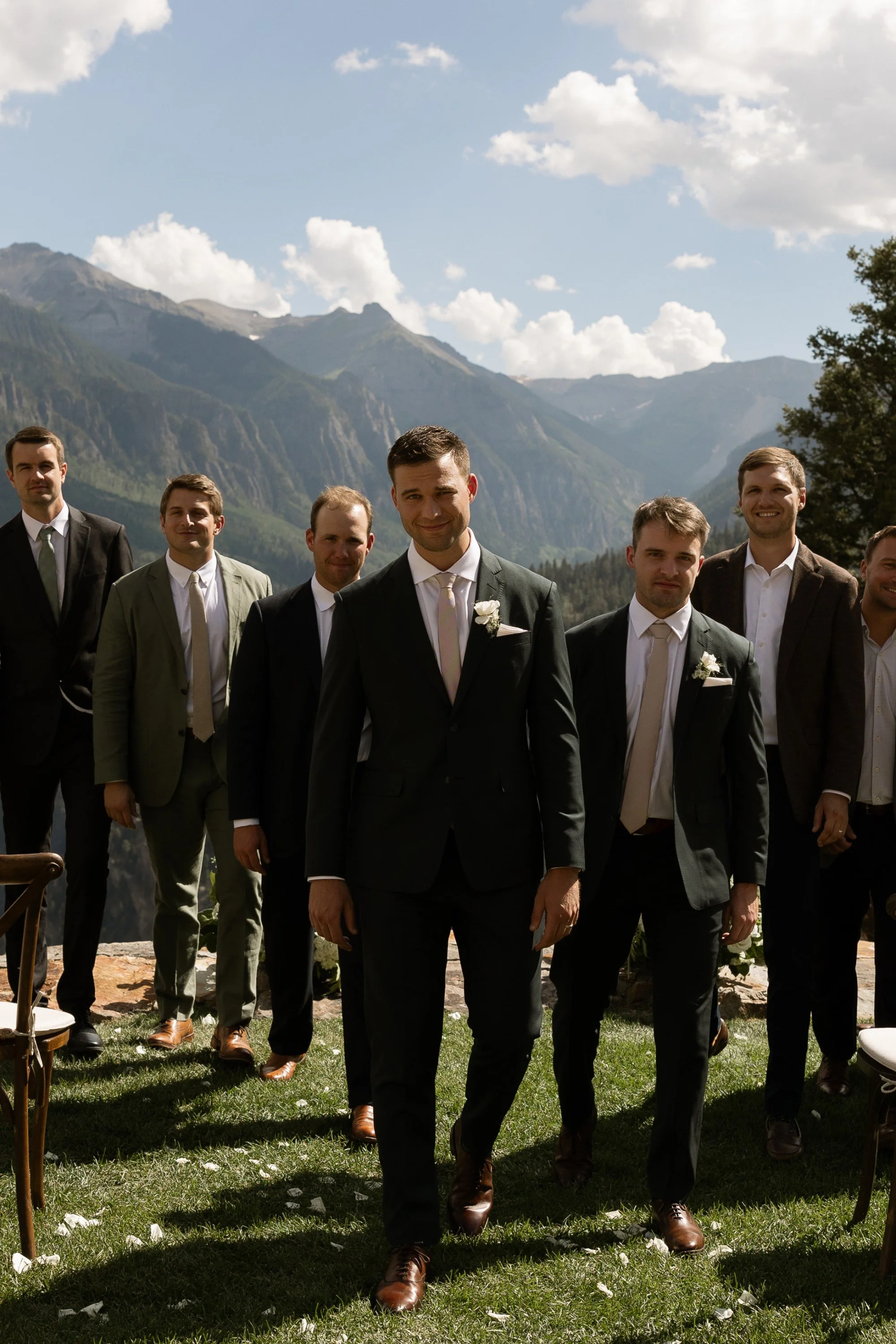 A group of men dressed in suits, walking outdoors during a wedding ceremony, with mountains and a cloudy sky in the background.
