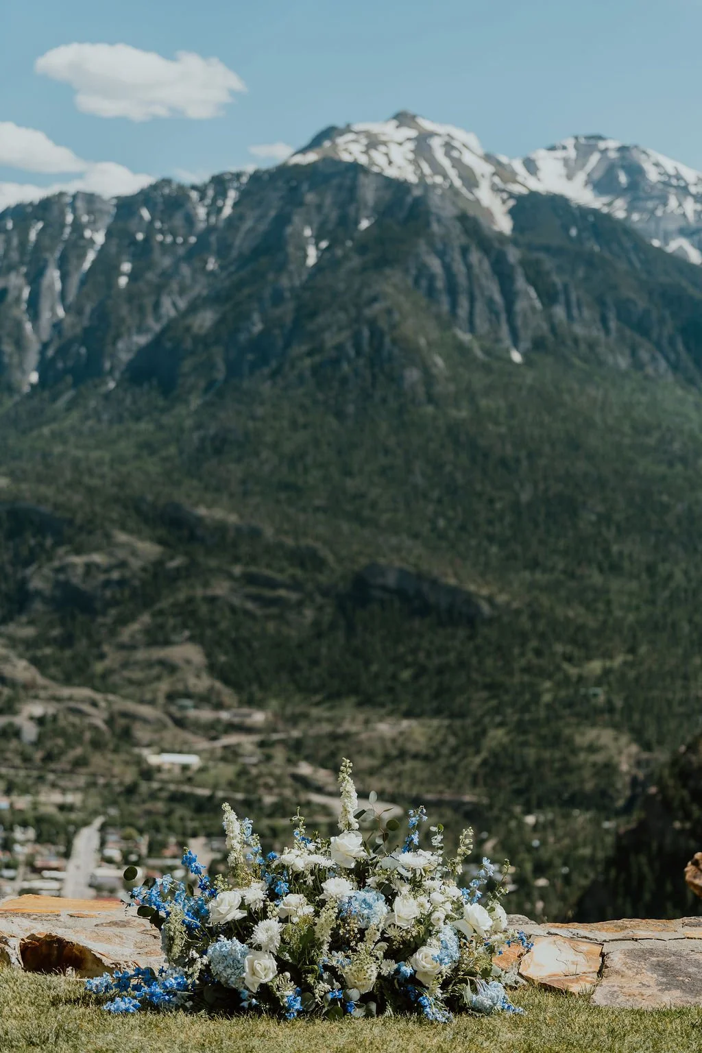 A bouquet of white and blue flowers placed on a stone ledge with mountains in the background.