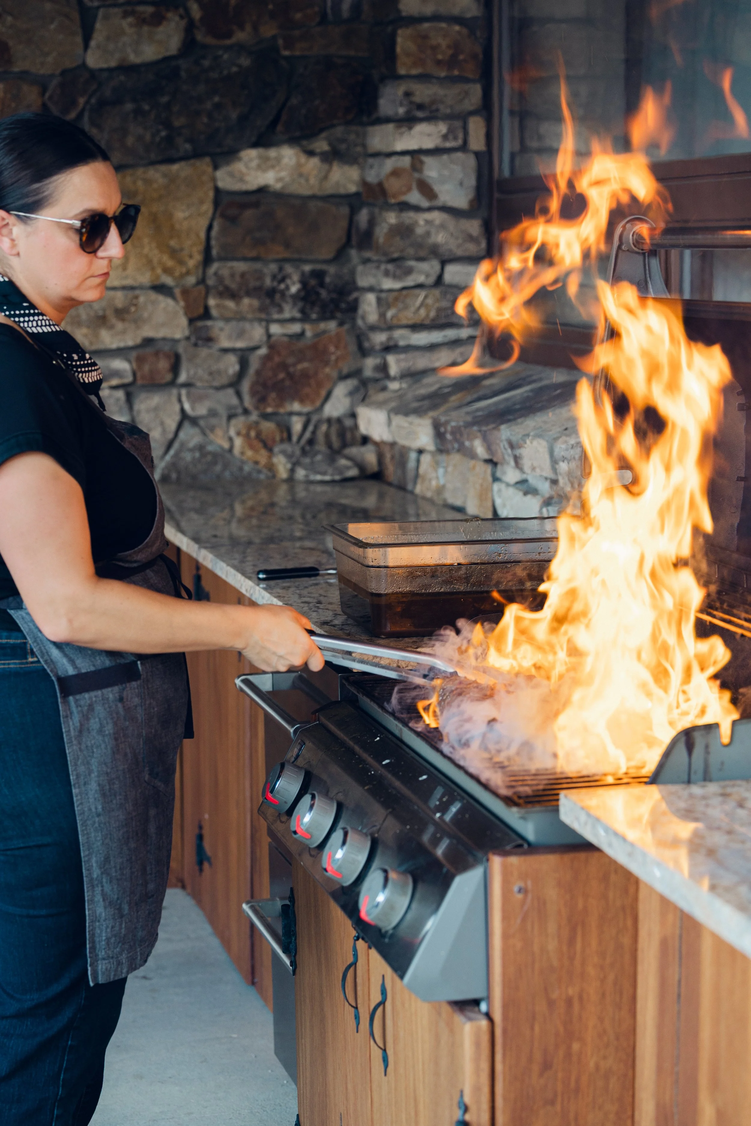 A woman wearing sunglasses and a gray apron struggles to put out a fire on the stove with a long utensil.