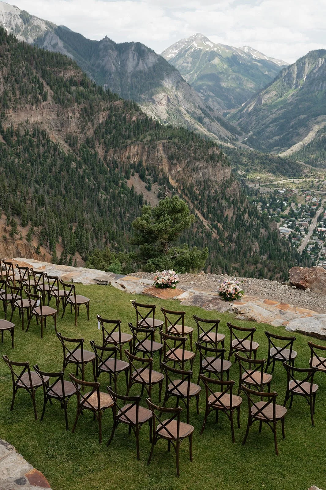 Empty chairs arranged outdoor for an event on a grassy area with mountain scenery in the background, including trees and snow-capped peaks.