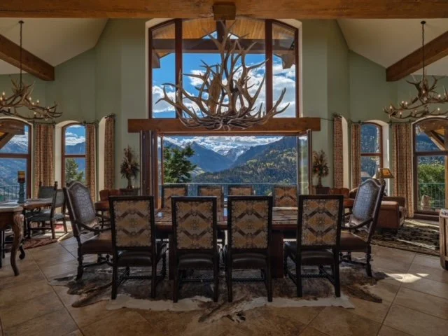 Interior of a dining room with large windows showing mountain views, antler chandelier, and rustic decor.
