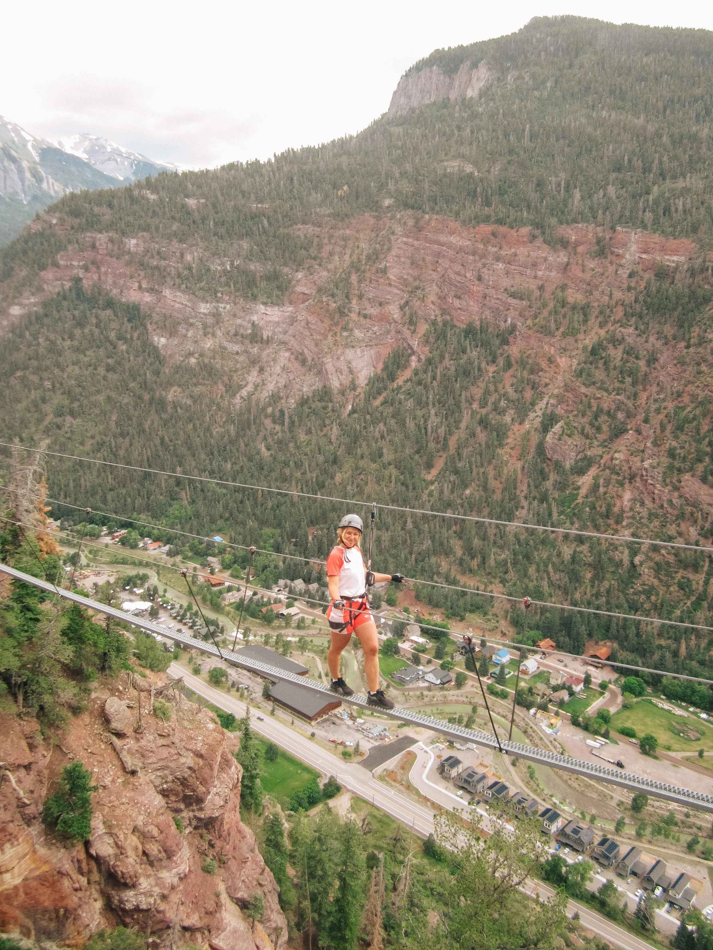 A woman on a safety harness walking across a suspension bridge high above a valley with mountains in the background.