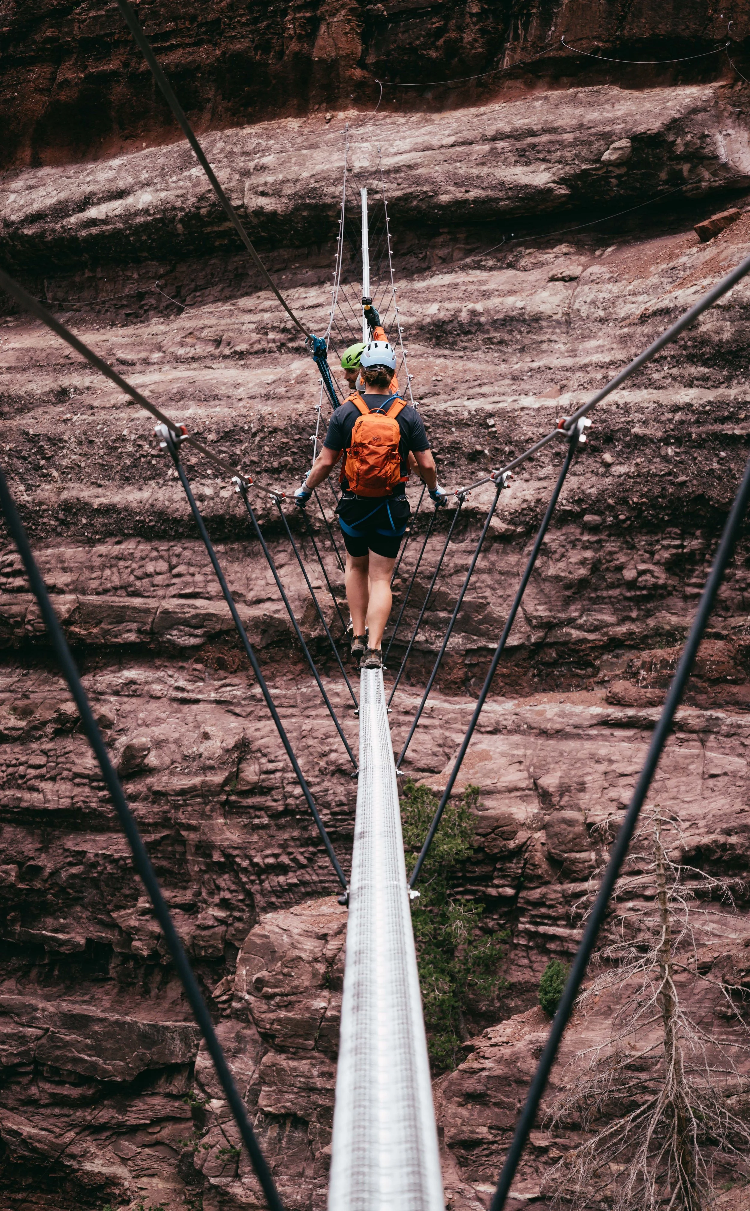 People walking across a suspension bridge over a rocky canyon, wearing helmets and backpacks.
