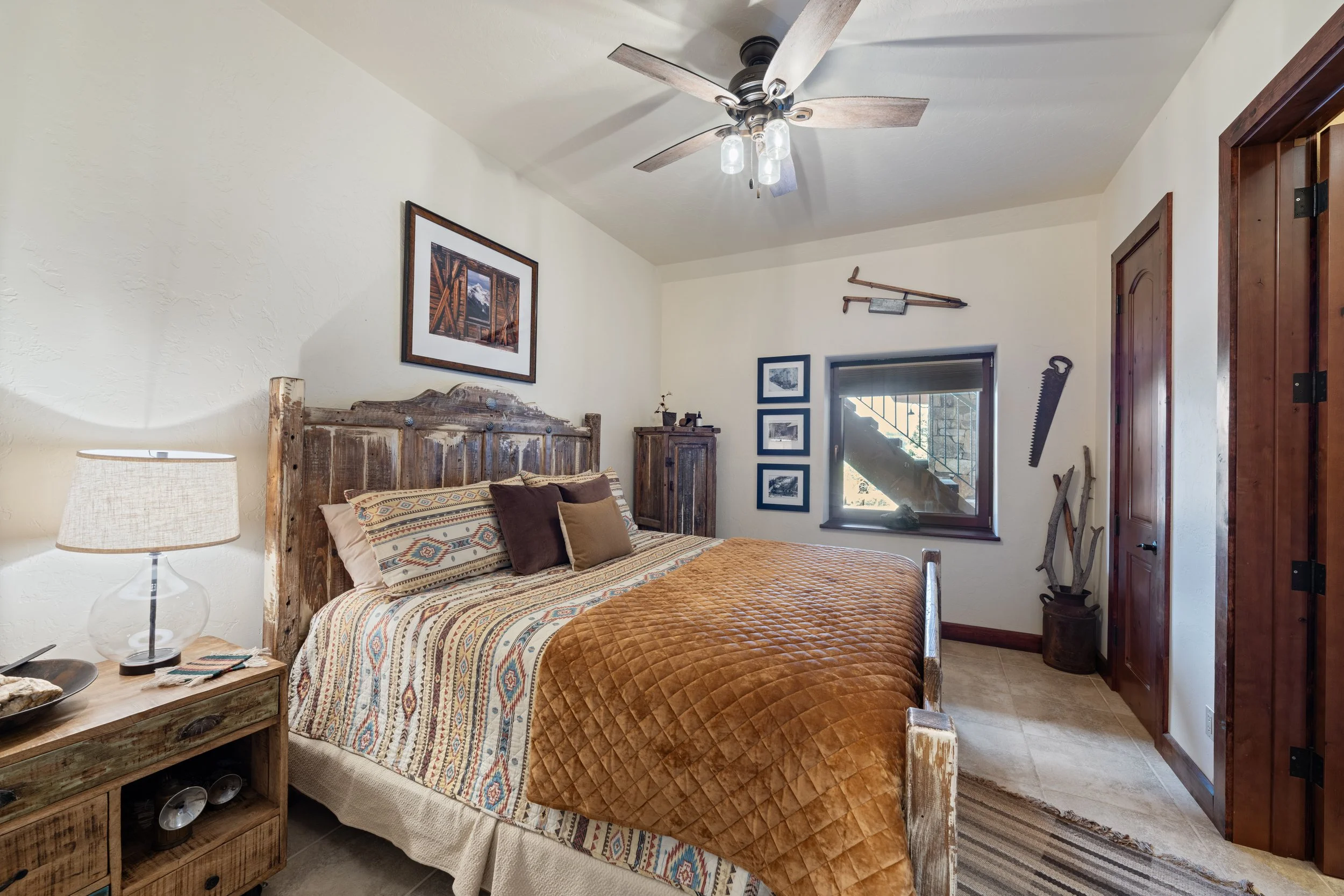 Cozy bedroom with rustic decor including a bed with patterned bedding and multiple pillows, a wooden nightstand with a lamp, framed mountain photographs and artwork on the walls, a ceiling fan, and vintage skis and saw wall decorations, with a small window revealing a staircase outside.