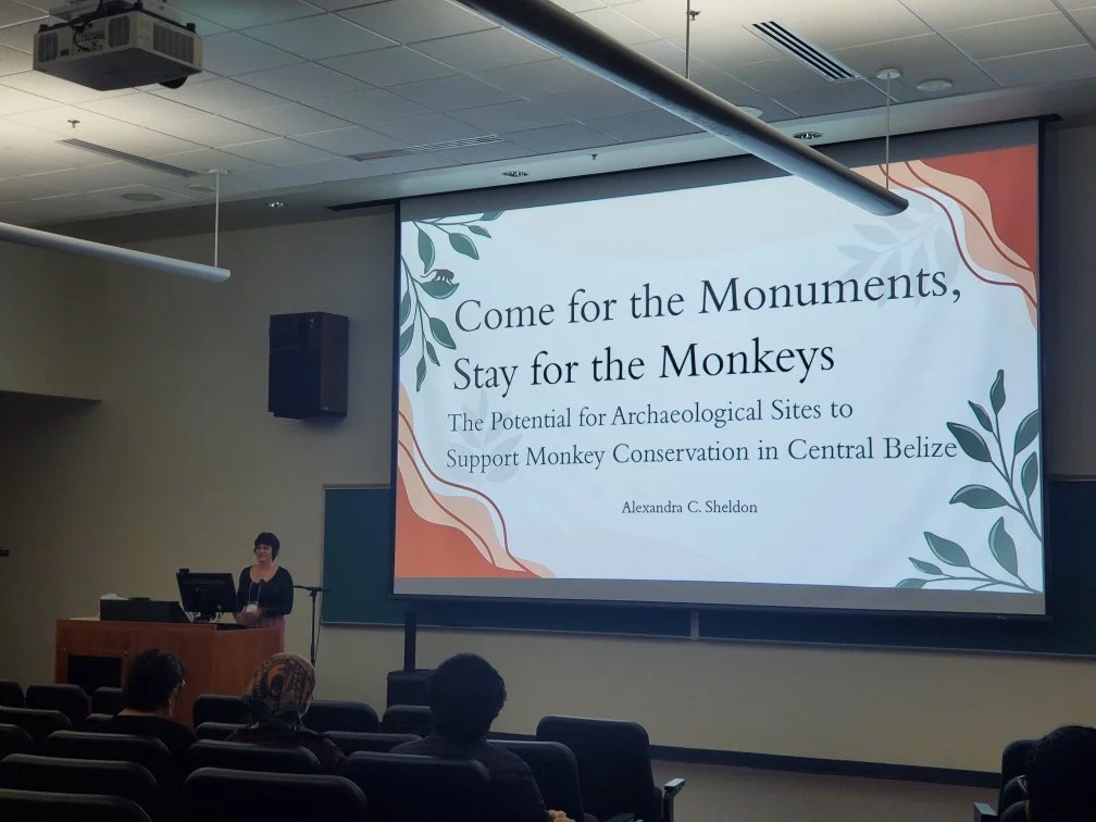 Woman standing behind a wooden podium with a screen projecting the title slide "Come for the monuments stay for the monkeys"
