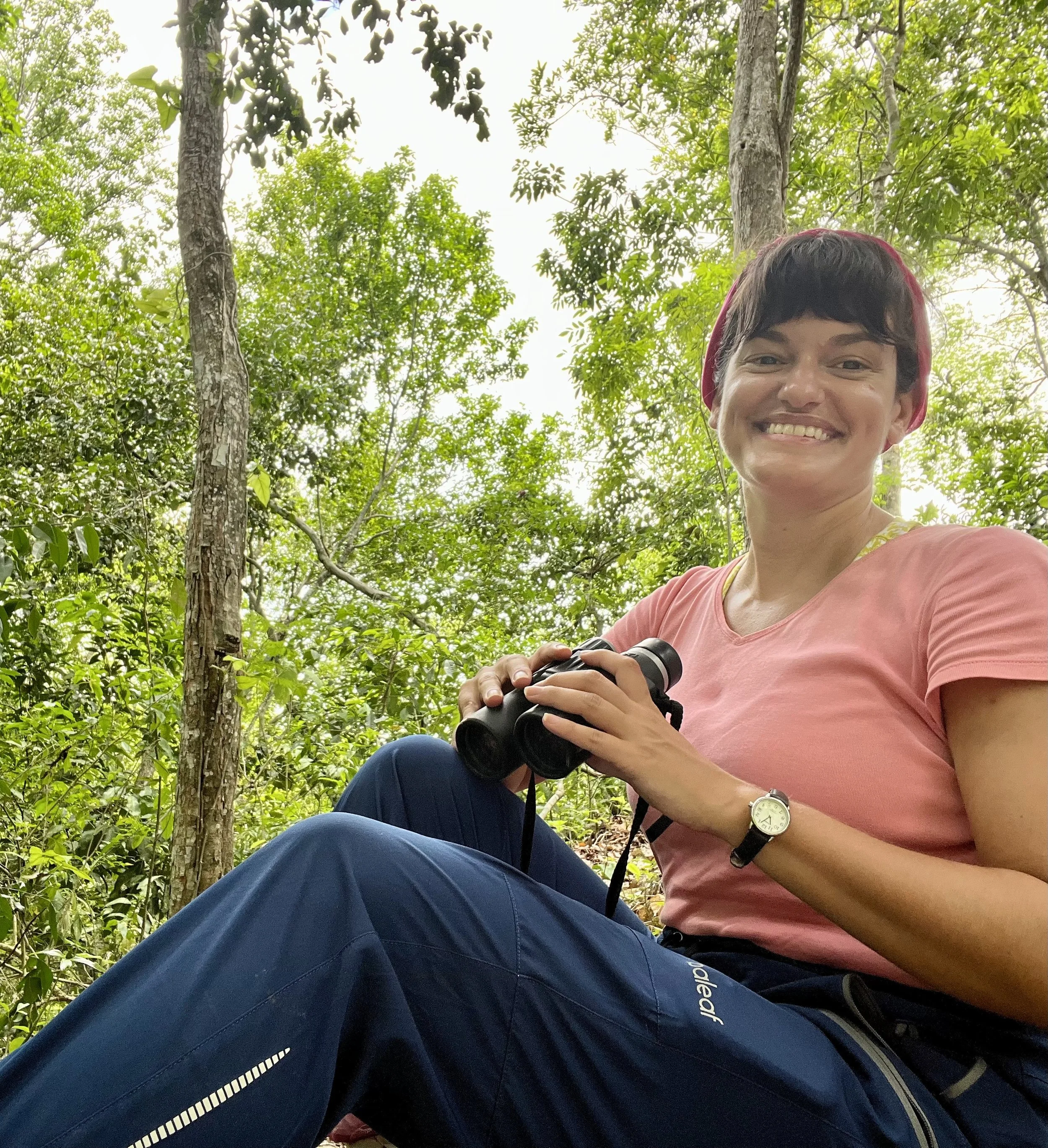 Female scientist smiles, holding binoculars and sitting on the ground in the forest