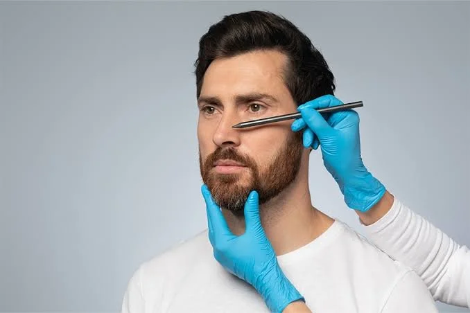 A man with a beard and mustache has a serious expression as a person in blue gloves examines his face with a metal tool, holding his chin and positioning the tool near his eye.