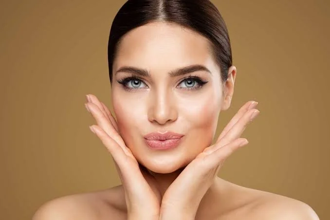 A woman with clear skin and dark brown hair styled straight, holding her hands near her face in a gentle pose, against a plain beige background.