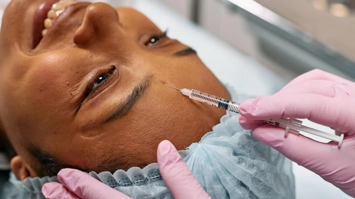 A person lying down receiving a cosmetic injection in their forehead from a medical professional wearing pink gloves.