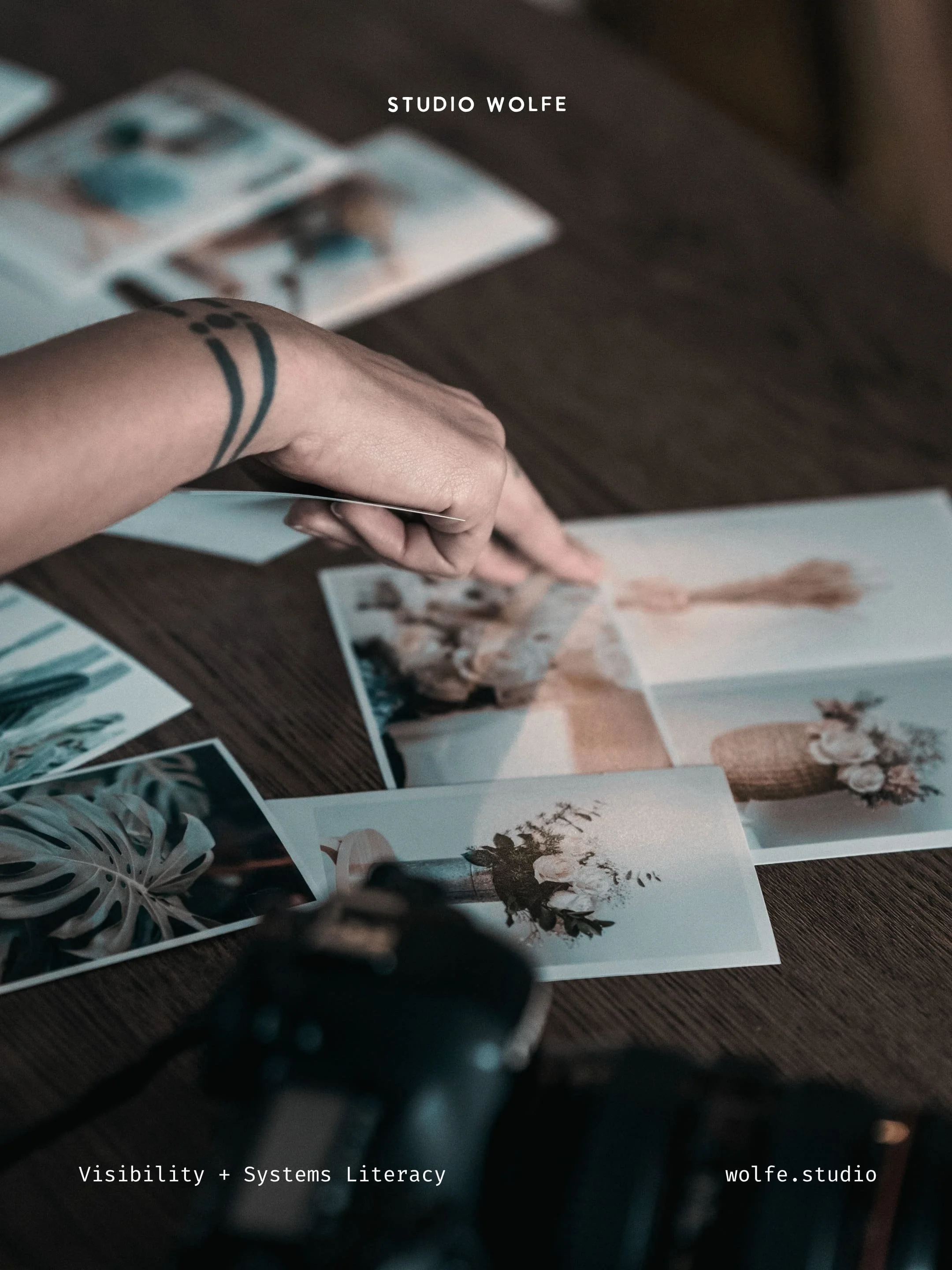 Hand selecting printed photographs on a wooden table beside a camera.
