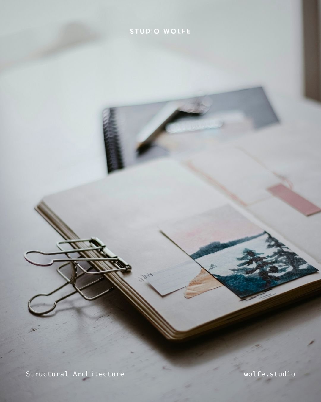 Open notebook with clipped visual references, a binder clip and soft-focus creative materials on a pale desk.