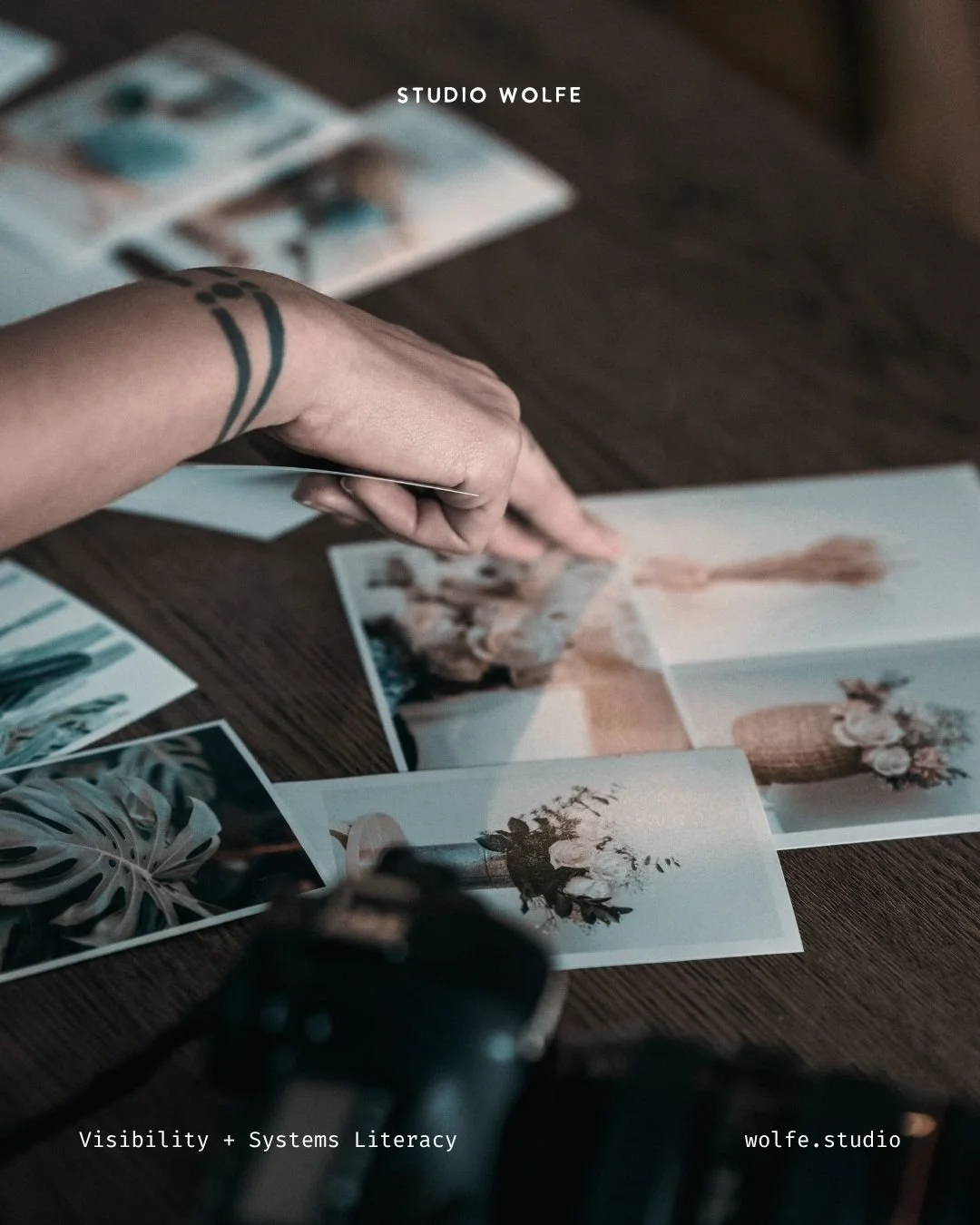 Hand selecting printed photographs on a wooden table beside a camera.