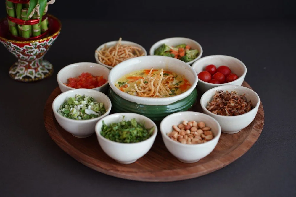 A wooden tray with nine small bowls. The central bowl contains soup with crispy noodles on top. Surrounding bowls hold fresh vegetables, chopped herbs, cherry tomatoes, peanuts, fried onions, and other garnishes.
