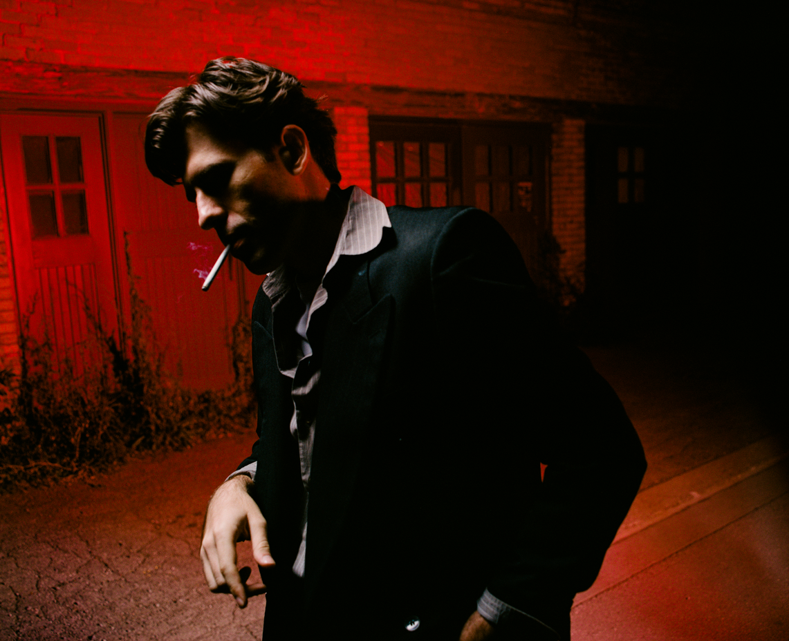 A young man with dark hair, wearing a dark suit and shirt, stands outdoors at night smoking a cigarette, with a red-toned background of a house and a driveway.